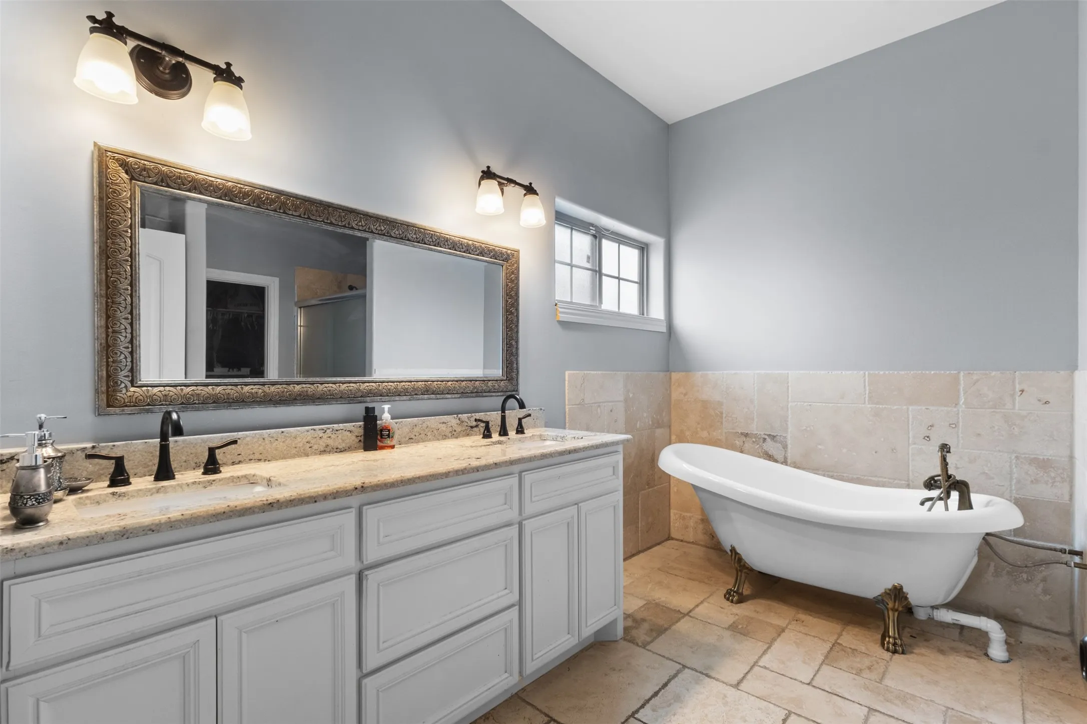 Dual vanity with granite countertop, standalone clawfoot tub, natural stone tile flooring, and a large framed mirror