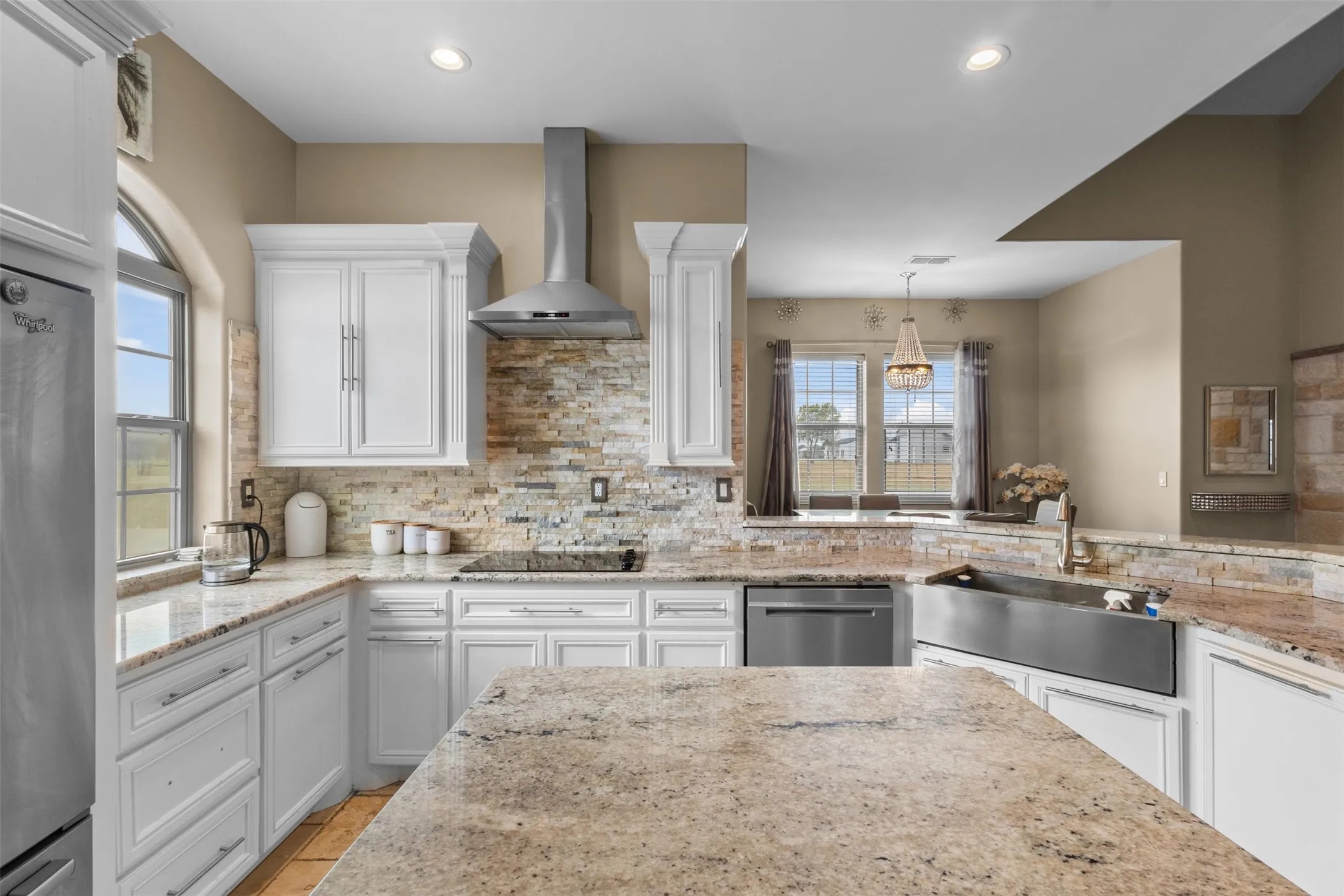 Spacious kitchen featuring white cabinetry, light-toned stone countertops, a stainless steel range hood, and a large stainless steel farmhouse sink