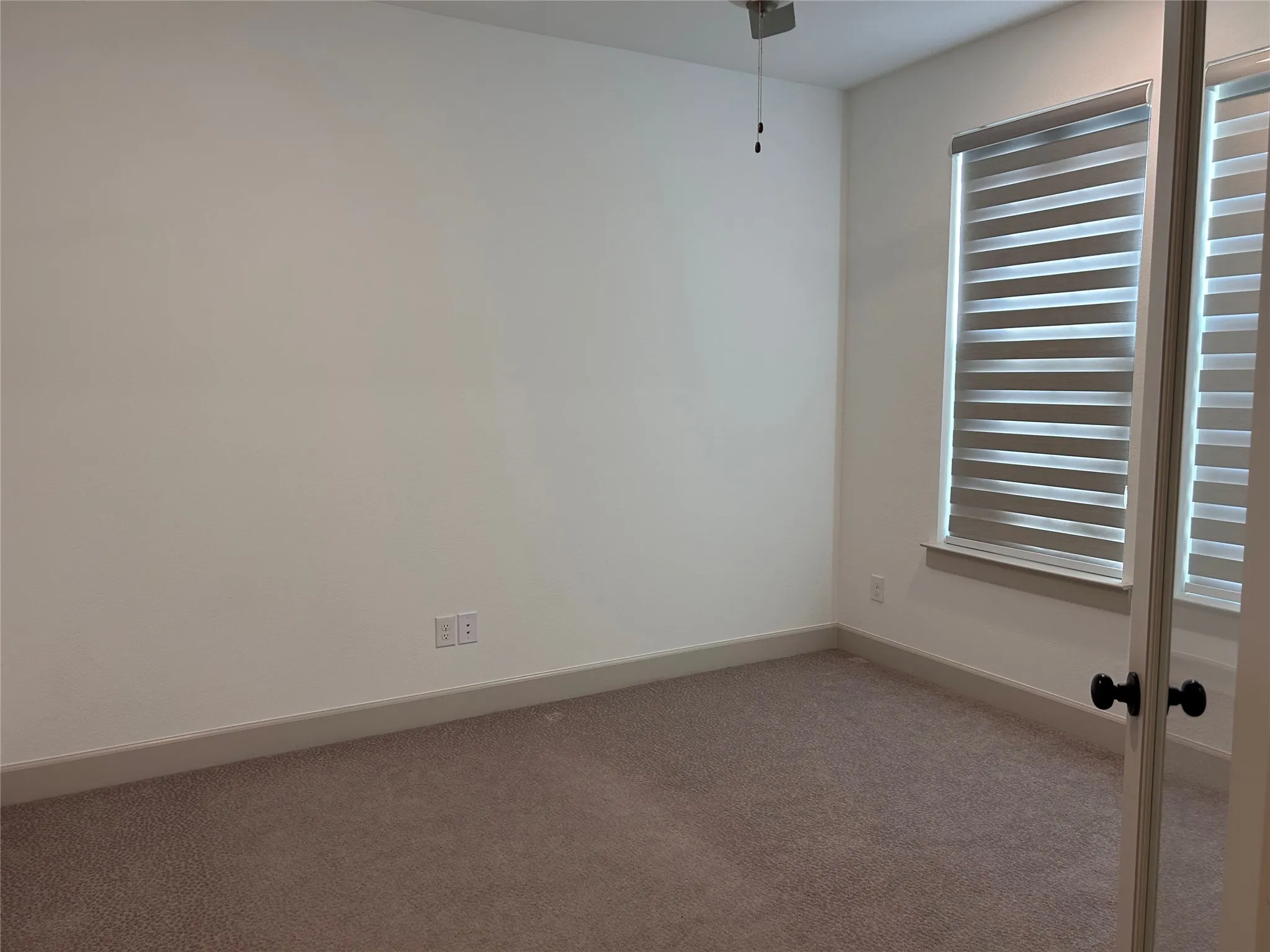 Neutral carpeting with white baseboards, a window with layered shades, and a ceiling fan with light fixture