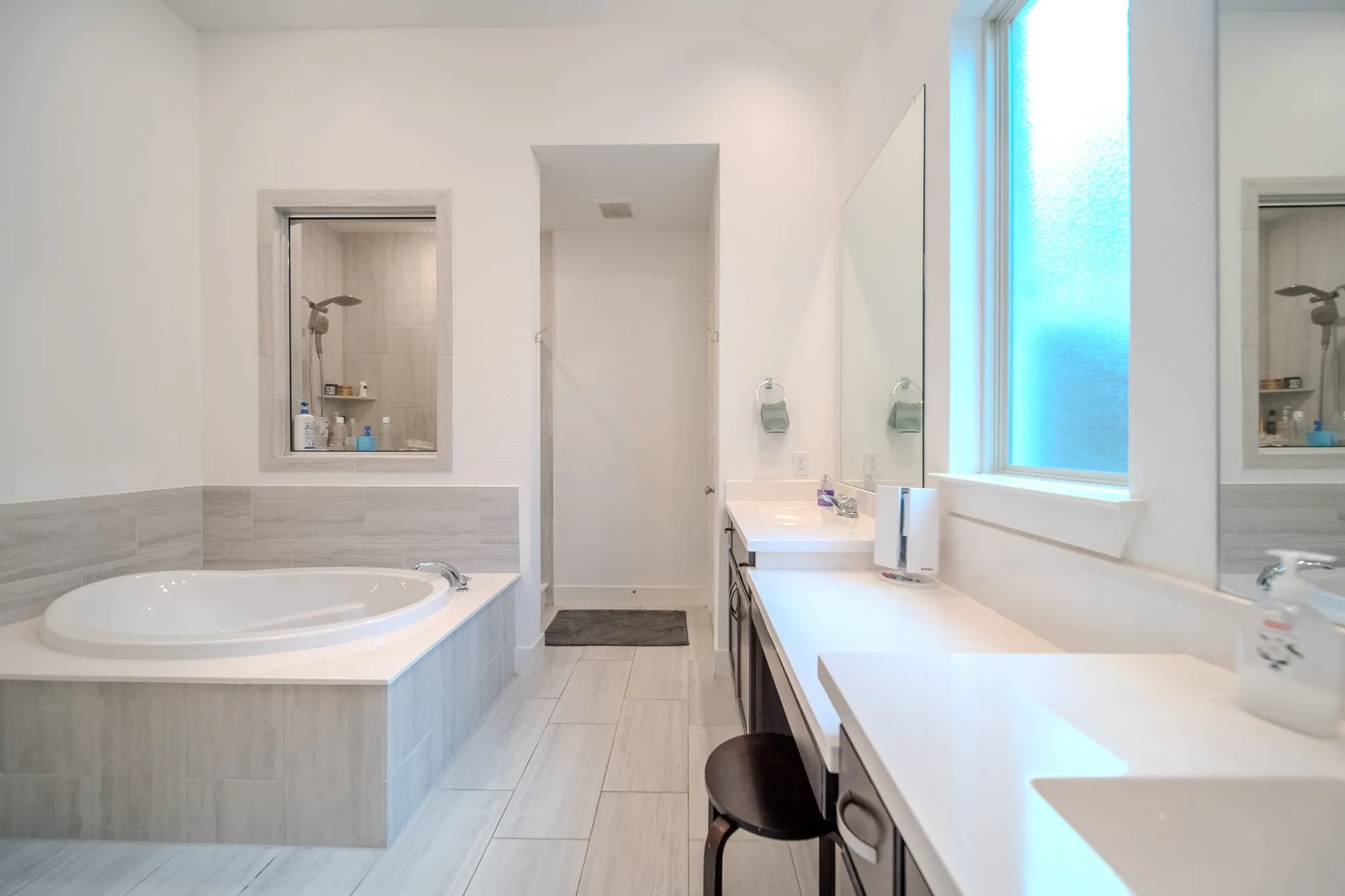 Built-in soaking tub with light-toned tile surround, dual vanity with undermount sinks, extended countertop space, and large privacy window