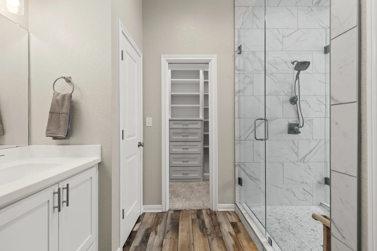 Glass-enclosed shower with subway tile and pebble flooring, wood-finish flooring, white vanity with integrated sink and quartz countertop, and a walk-in closet with built-in shelving and drawers