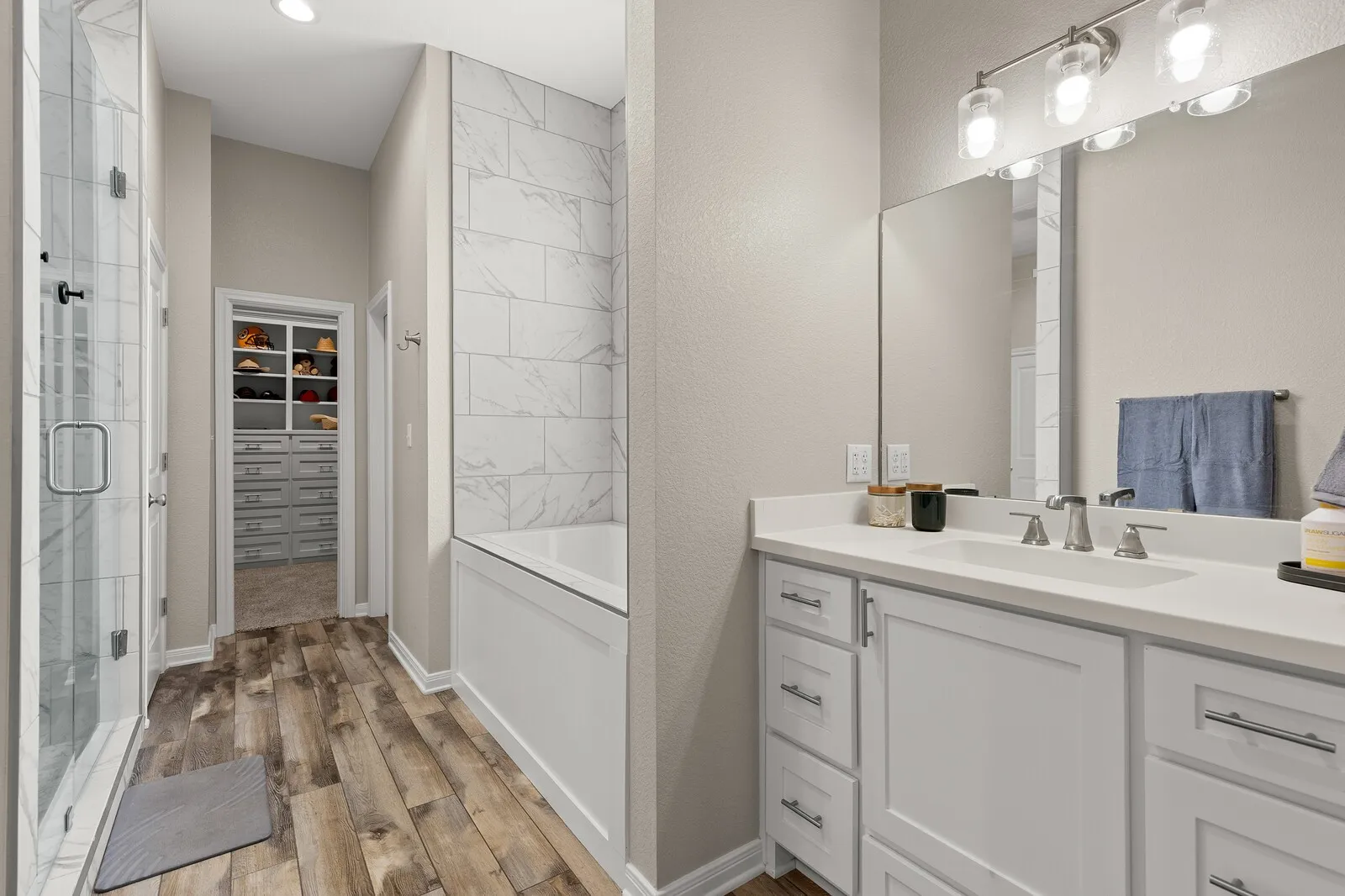 Dual vanity with white cabinetry and chrome hardware, glass-enclosed shower with tile surround, built-in bathtub with subway tile accent wall, wood-finish flooring, and a walk-in closet with built-in shelving