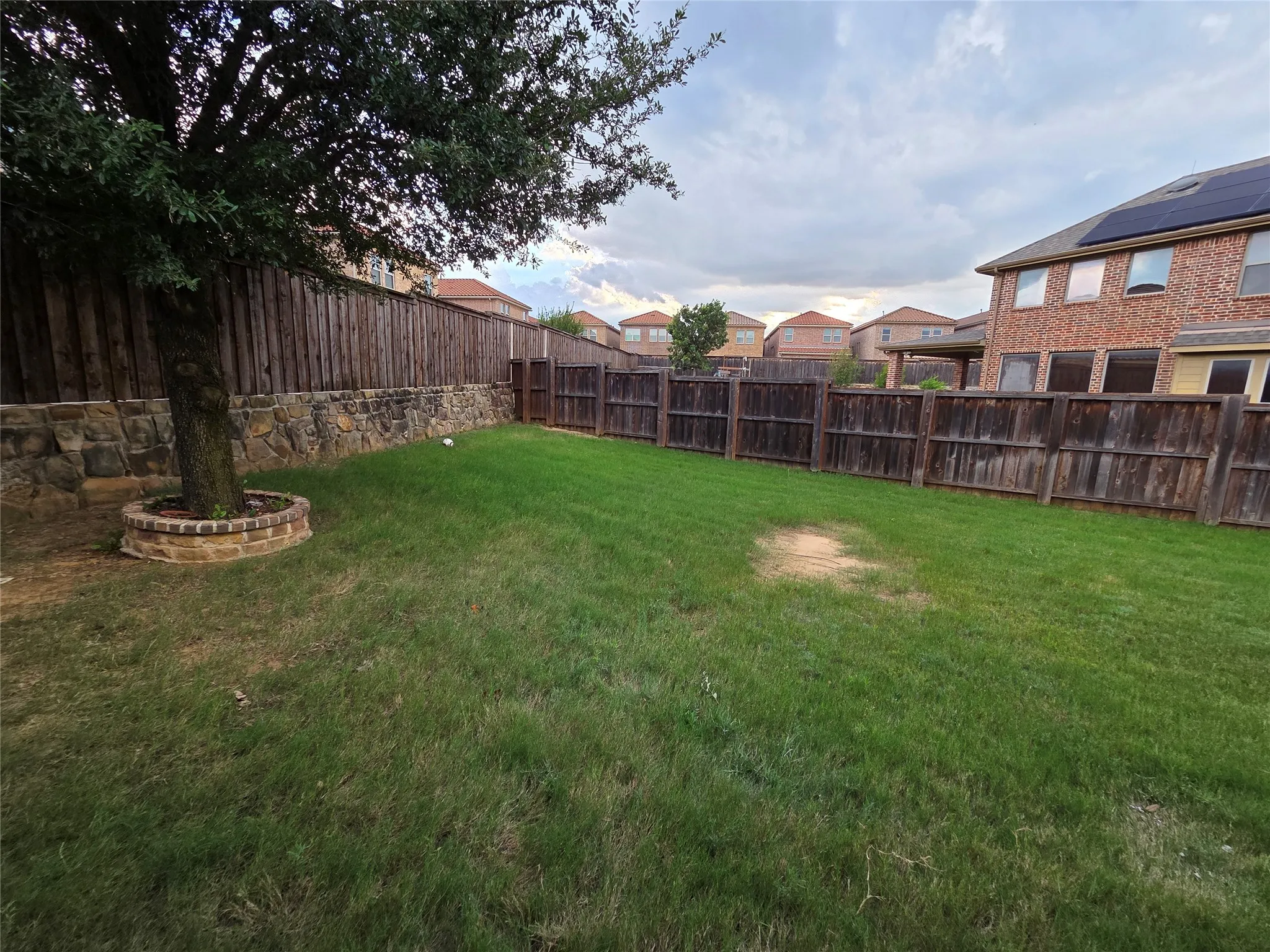 Expansive backyard featuring a manicured lawn, established tree, and a stone retaining wall with brick accents