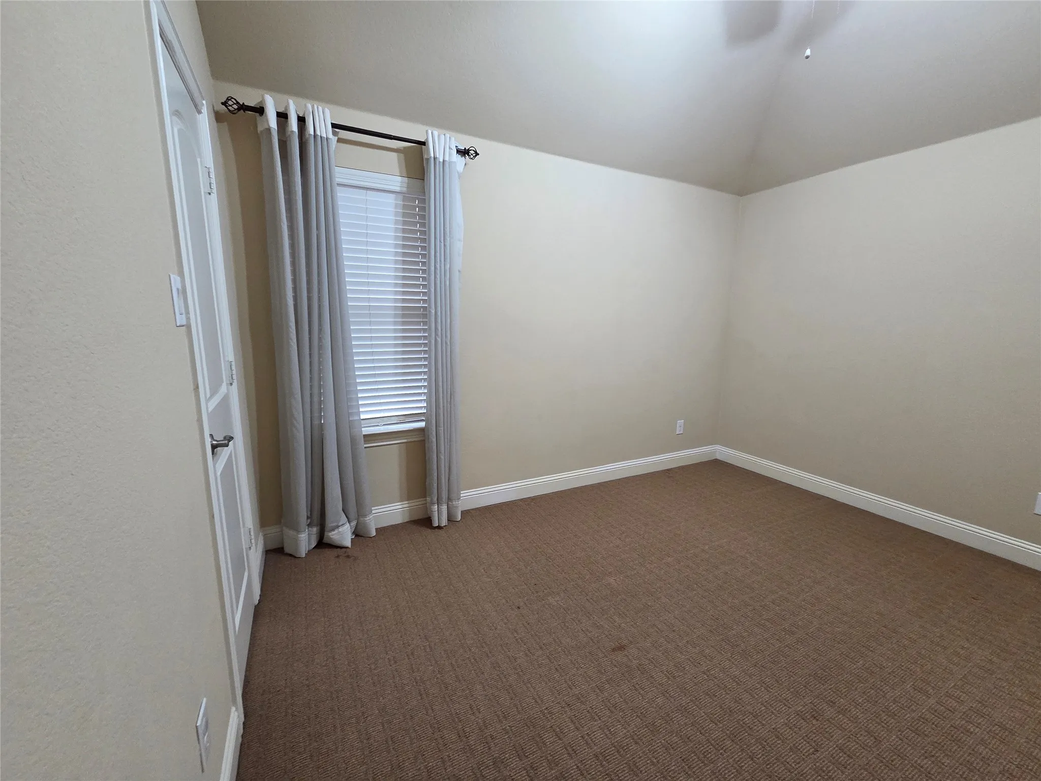 Carpeted flooring with light-toned walls, a single window with blinds and drapes, and a vaulted ceiling