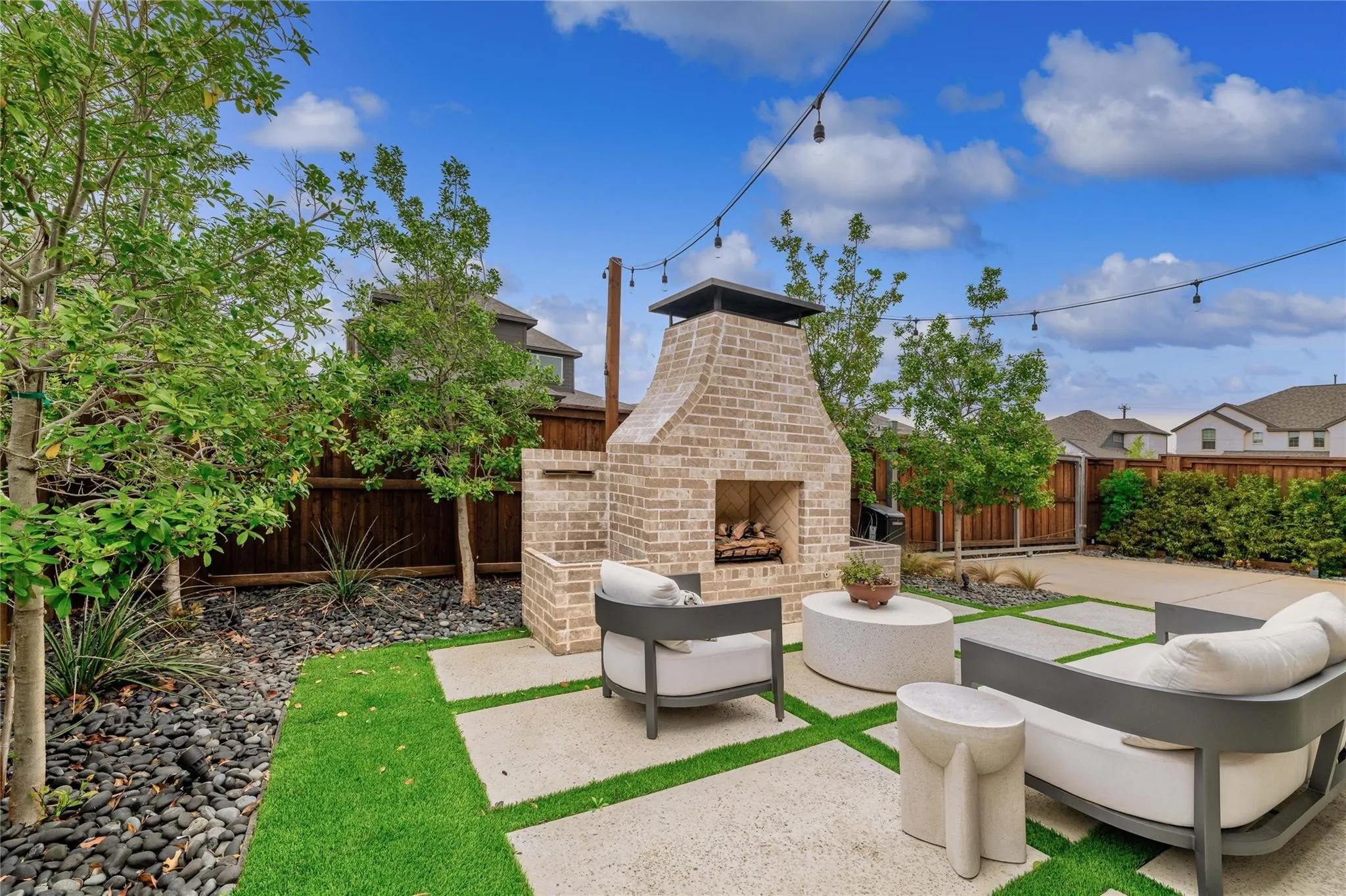 Landscaped backyard featuring a brick outdoor fireplace, string lighting, concrete pavers, artificial turf sections, and a dark wood fence
