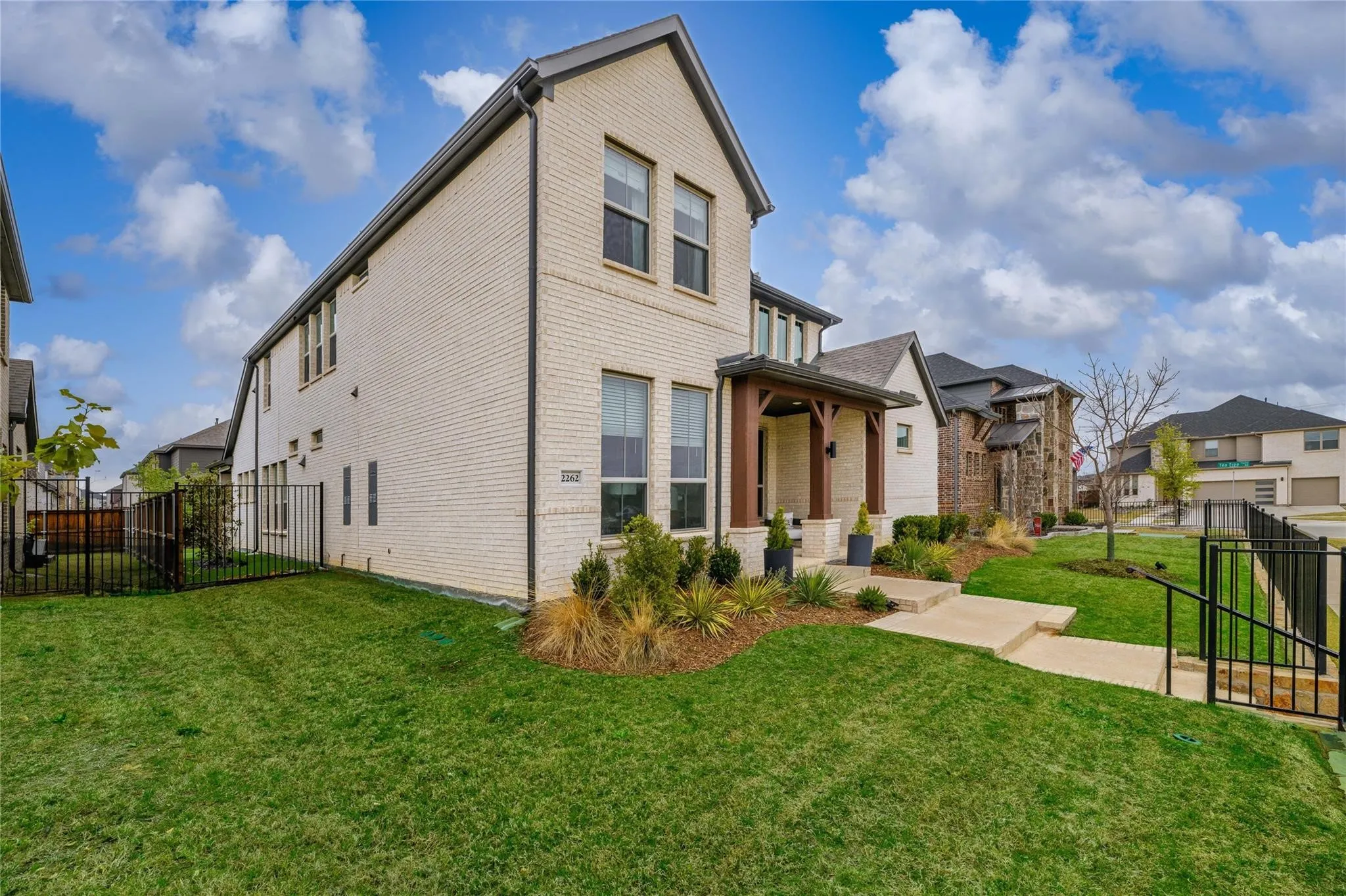 Brick facade with dark trim, covered entry with wood columns, front yard landscaping, and a black metal fence