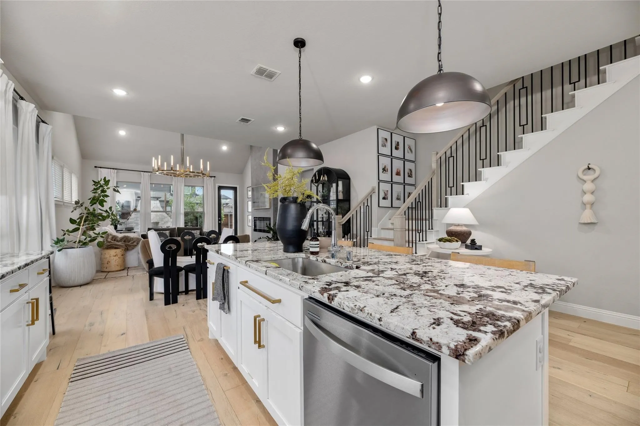 Open-concept living area featuring a kitchen island with stone countertop, stainless steel appliances, light wood-finish flooring, and a modern staircase with dark metal balusters
