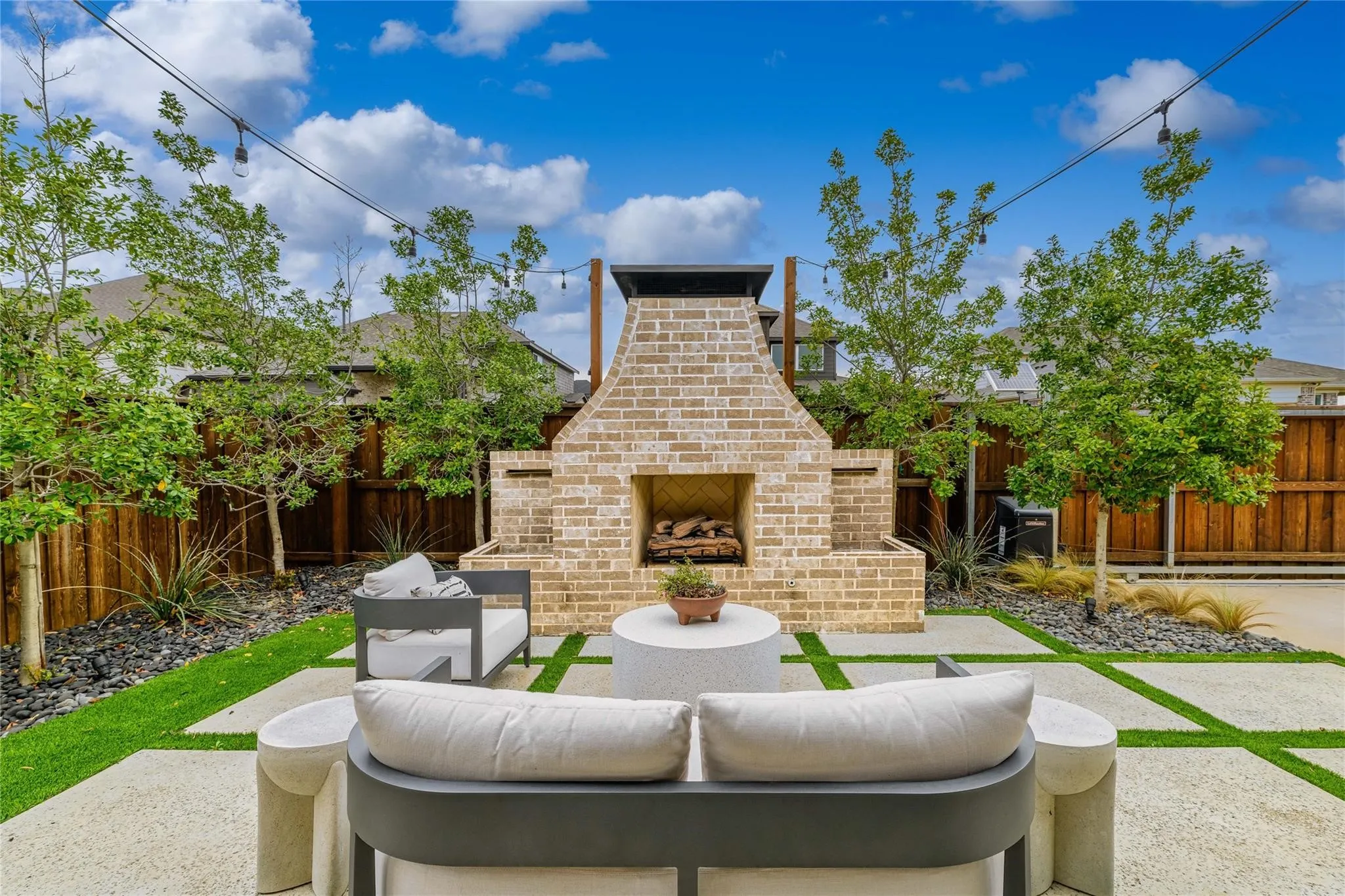 Expansive outdoor living area featuring a brick fireplace, integrated wood storage, string lighting, and a combination of concrete pavers with artificial turf