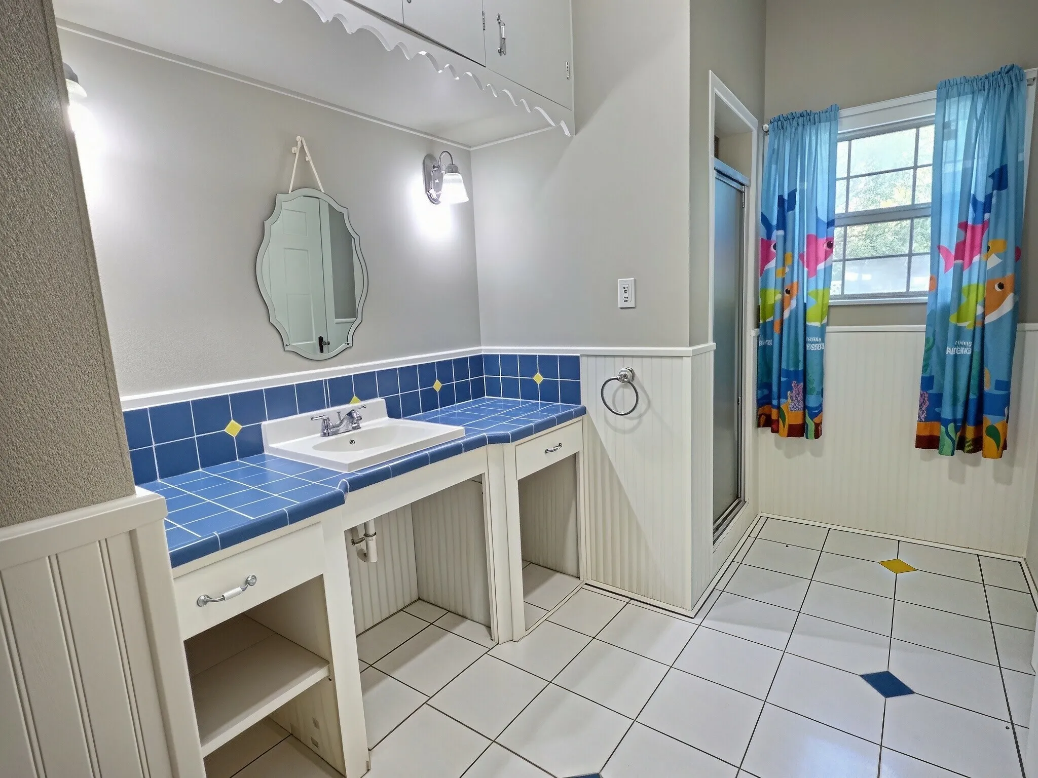 Vanity with tile countertop and integrated sink, white beadboard wainscoting, white tile flooring, and a glass-enclosed shower