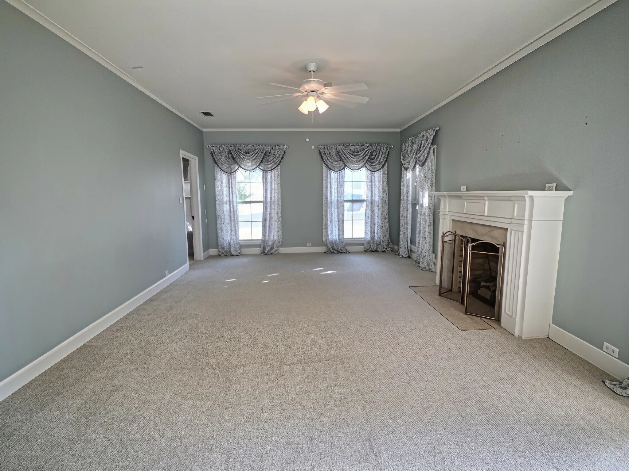 Spacious room featuring a decorative fireplace with a mantel, multiple windows, wall-to-wall carpeting, and a ceiling fan