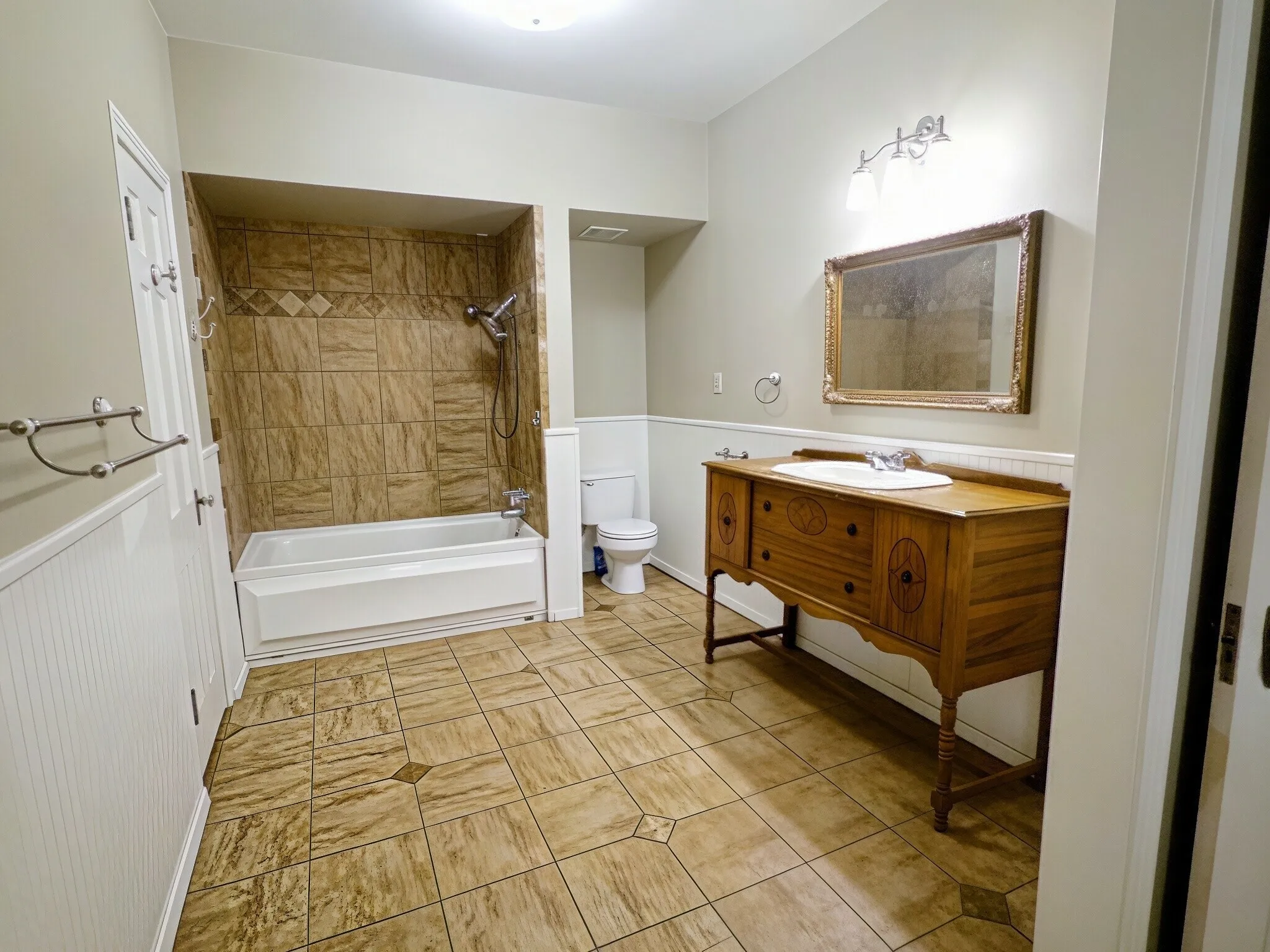 Tiled flooring with accent patterns, wood-finish vanity with an integrated sink, shower-tub combination with full wall tiling, and beadboard wainscoting