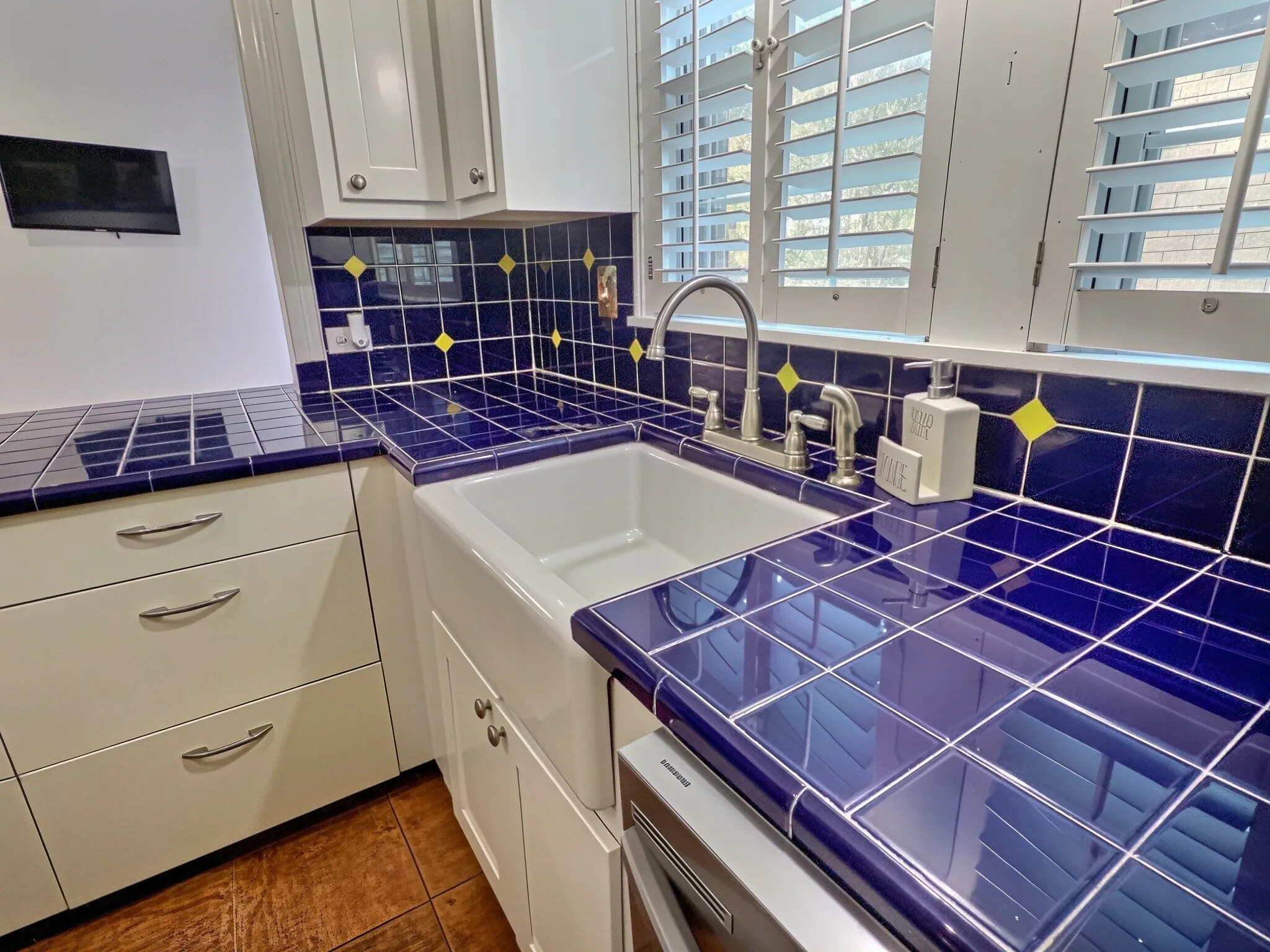 White cabinetry with brushed nickel hardware, blue and yellow tiled backsplash, farmhouse-style sink with gooseneck faucet, window with white plantation shutters, and tiled countertops