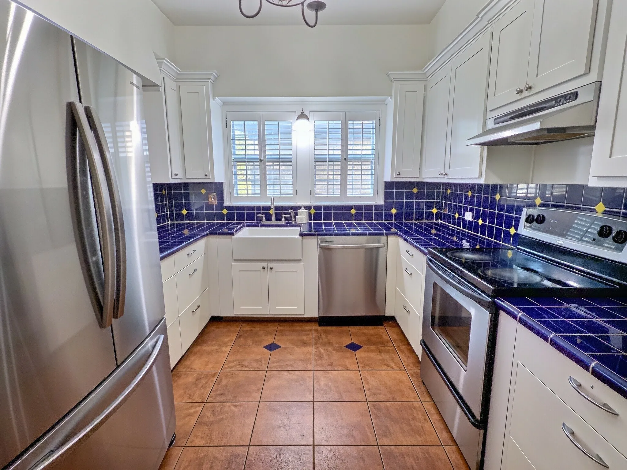Kitchen featuring white shaker cabinetry, stainless steel appliances, a farmhouse sink, blue tile countertops, and terra cotta tile flooring