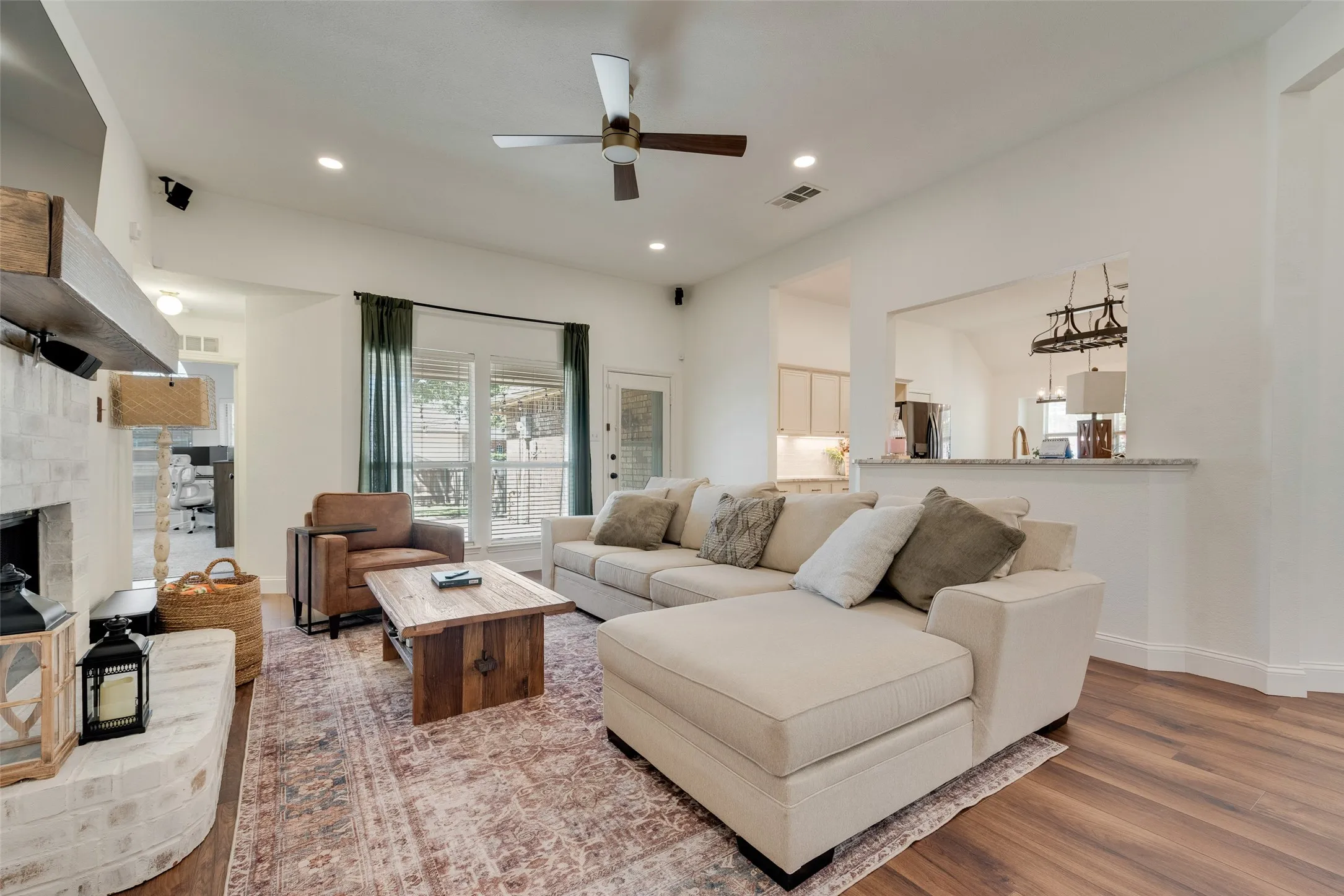 Vaulted ceiling with a ceiling fan, wood-finish flooring, brick-faced fireplace with a wood mantel, and a pass-through opening to the kitchen