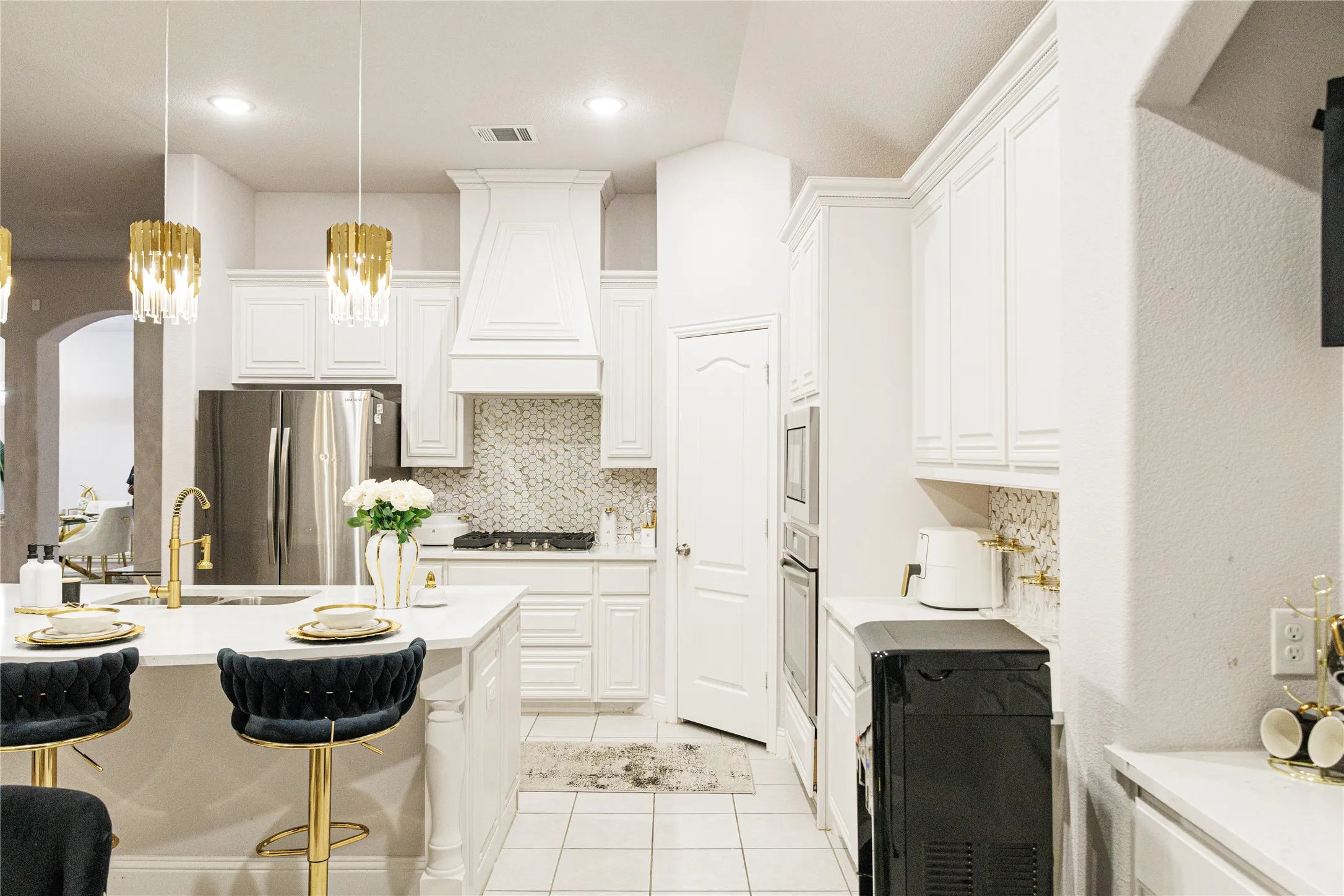 White cabinetry with crown molding, stainless steel refrigerator, built-in double oven, decorative range hood, and white tile flooring