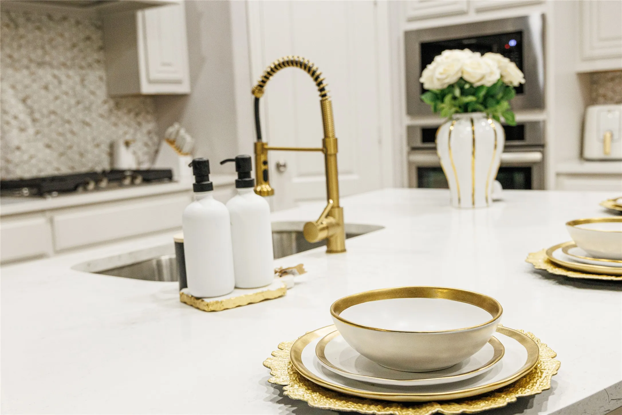 White countertop island with an integrated sink, gold-finish faucet, and stainless steel built-in double oven