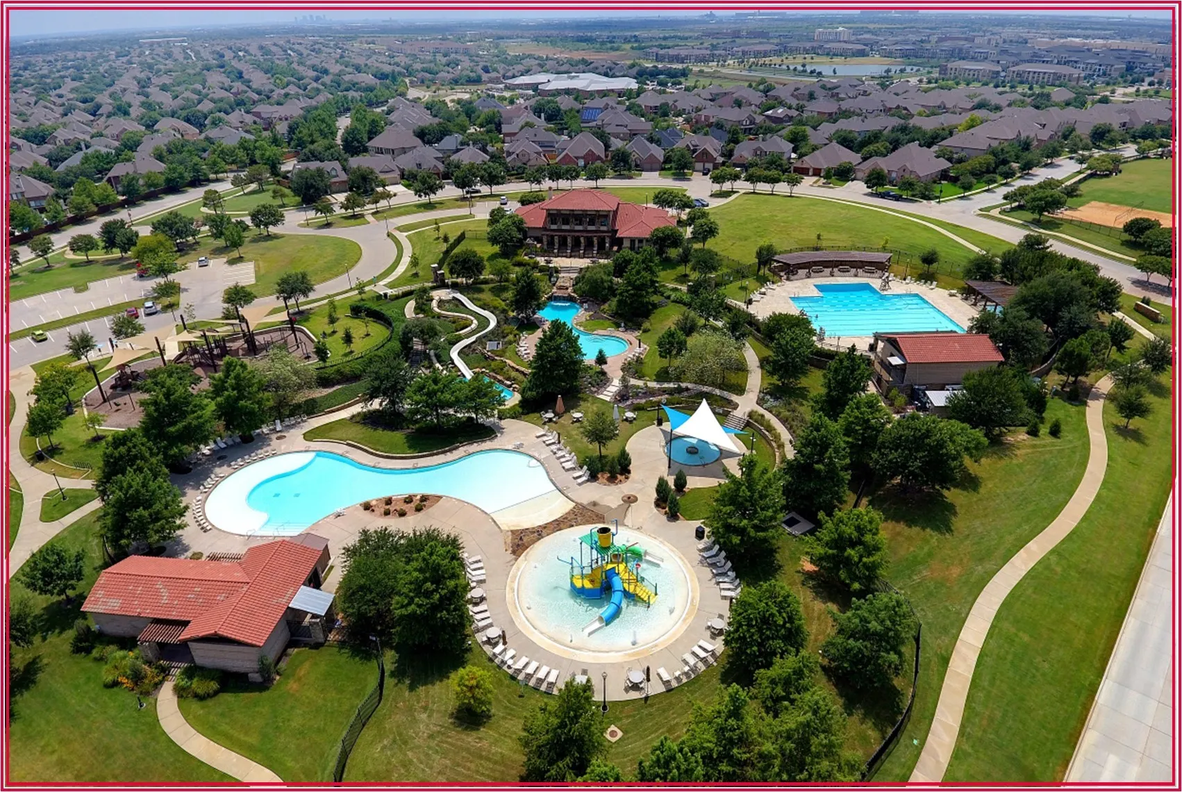 Community recreation area featuring multiple swimming pools, a water play structure, extensive green spaces, and paved pathways