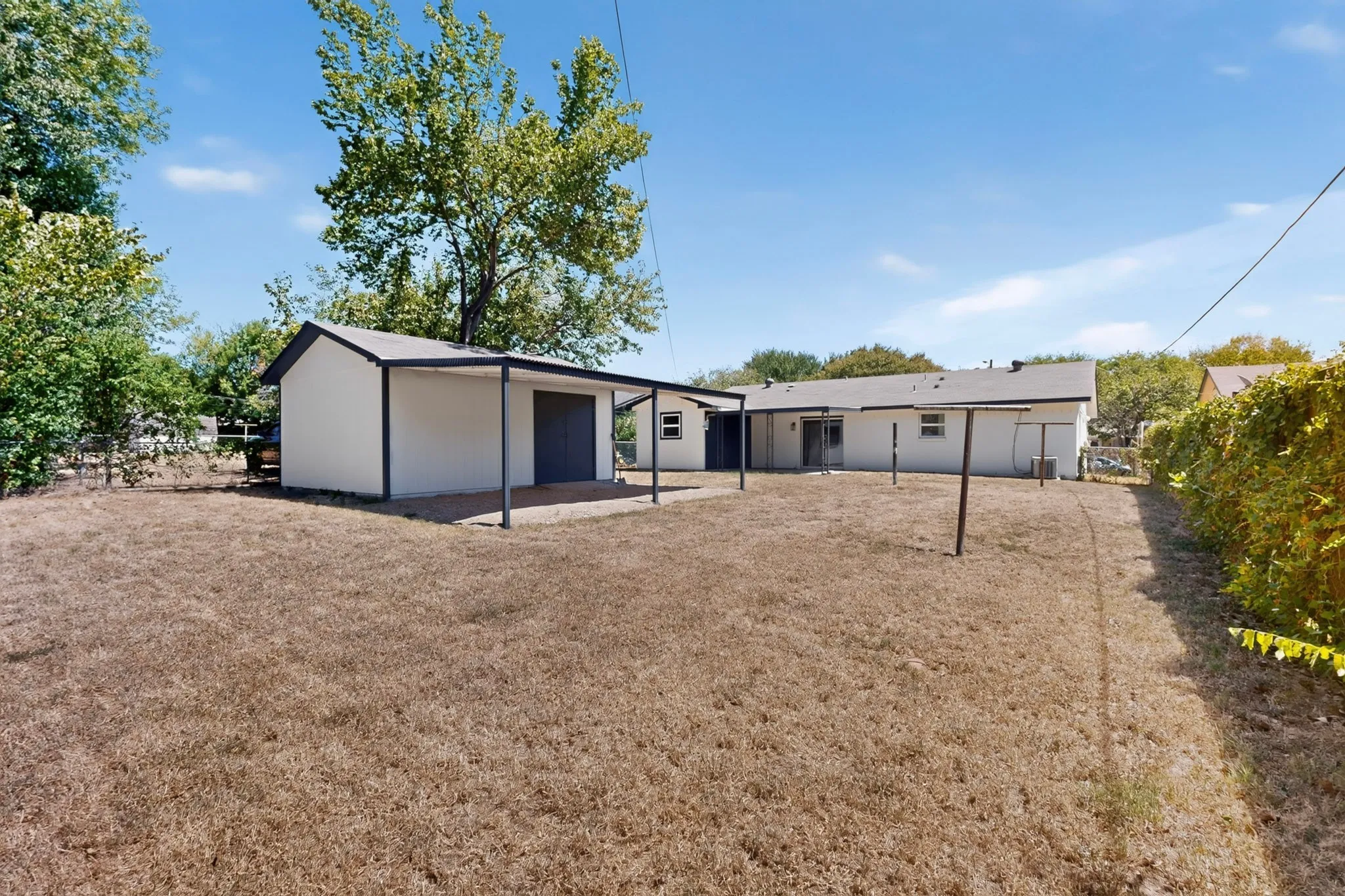 Expansive rear yard featuring a detached auxiliary structure with an attached carport, a covered patio, and mature perimeter trees