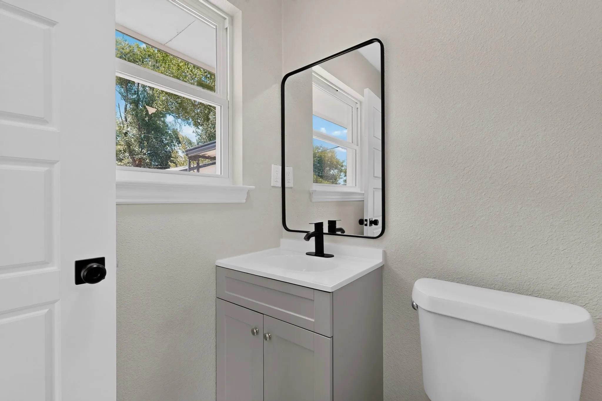 White paneled door with black hardware, window with white trim, light gray vanity with white countertop, black faucet, and frameless mirror
