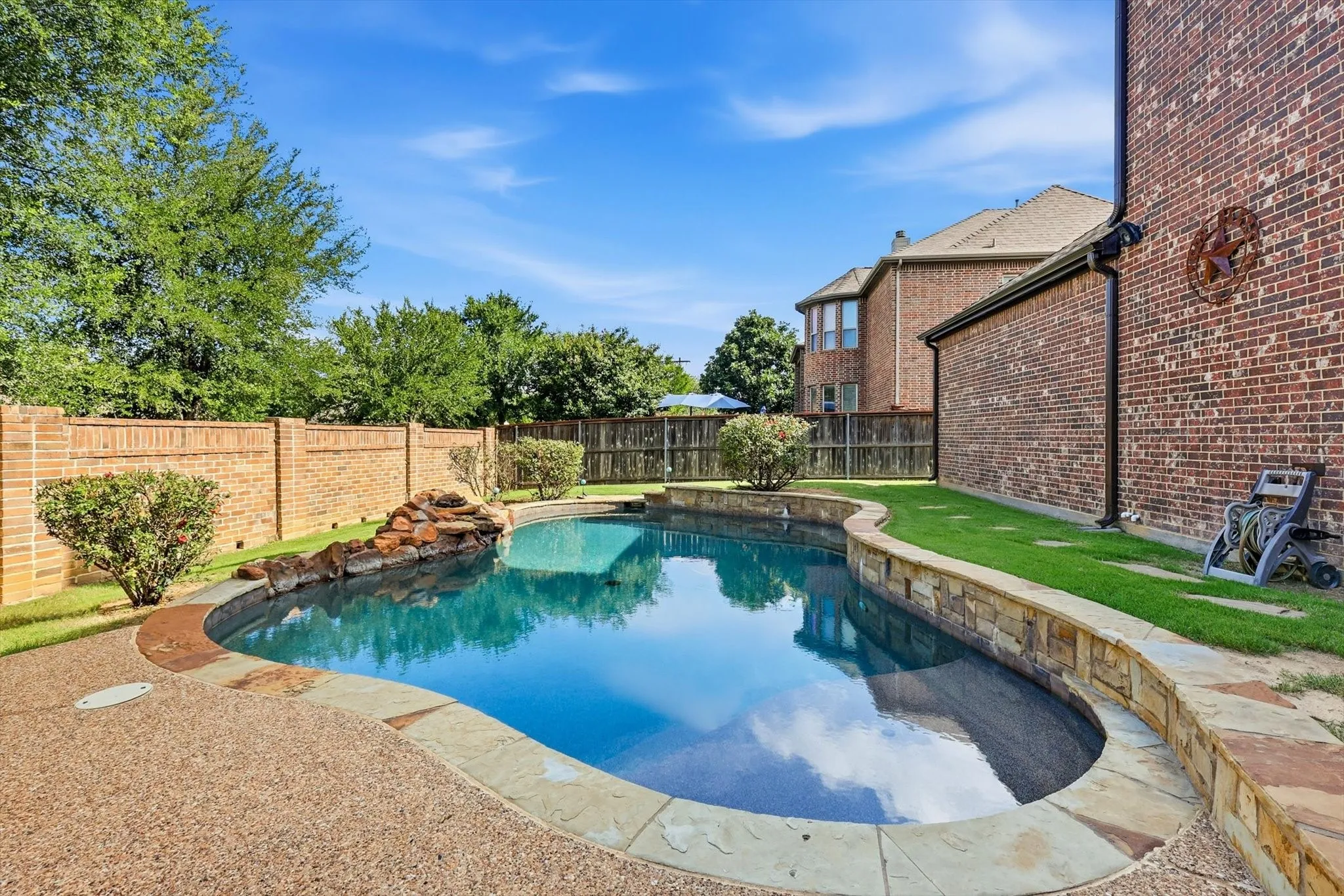Curved swimming pool featuring a rock waterfall, stone coping, and exposed aggregate decking