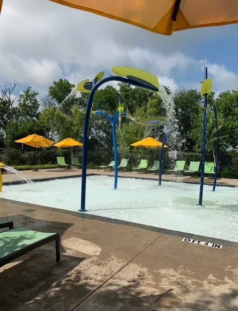 Splash pad recreation area featuring a zero-entry water feature with interactive spray elements, curved blue archway structure, and yellow umbrellas providing shade