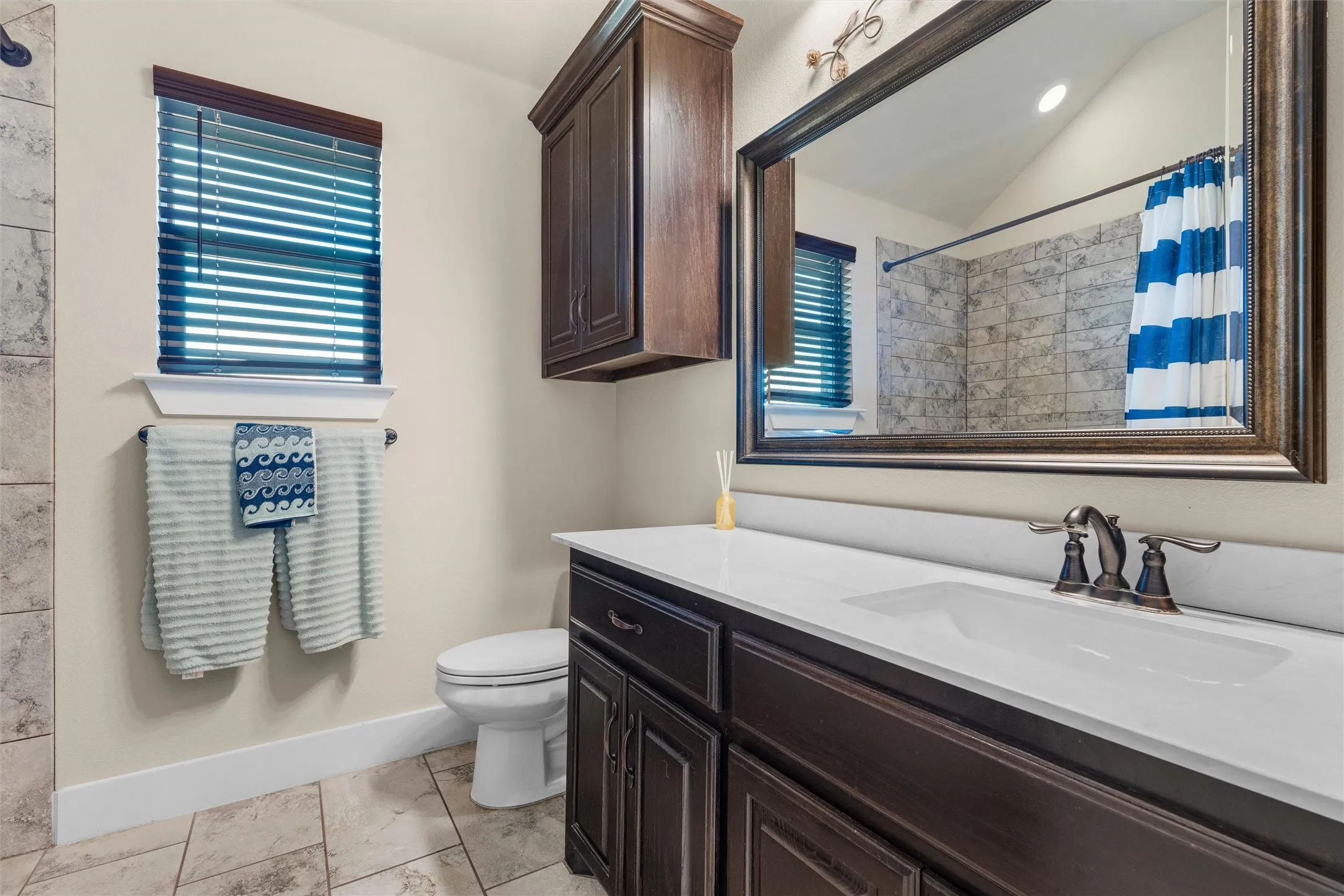 Custom vanity with integrated sink and dark bronze hardware, framed mirror, tiled flooring, built-in upper cabinetry, and a window with blinds