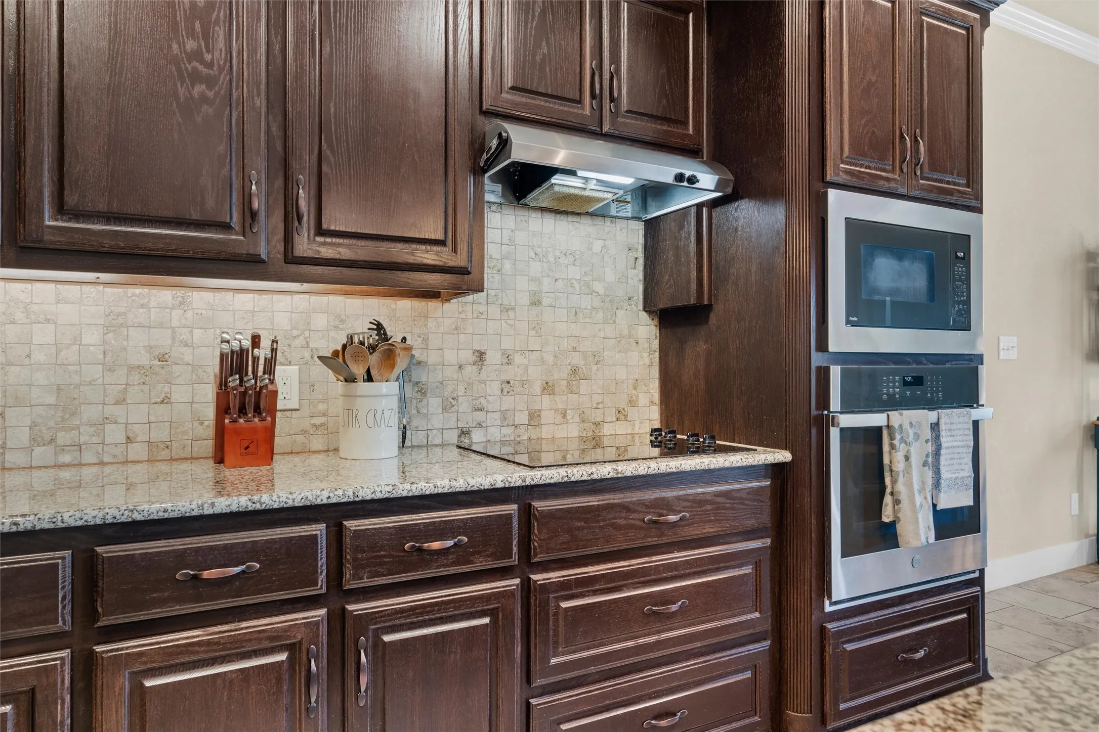 Dark wood cabinetry, granite-style countertops, mosaic tile backsplash, built-in stainless steel double oven, and a built-in stainless steel microwave