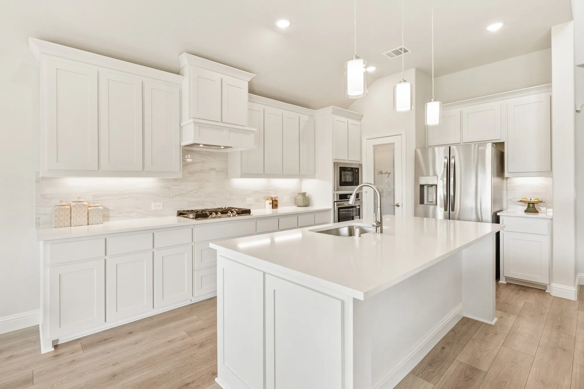 White shaker-style cabinetry, stainless steel appliances, light-toned wood-finish flooring, white countertops, and contemporary pendant lighting