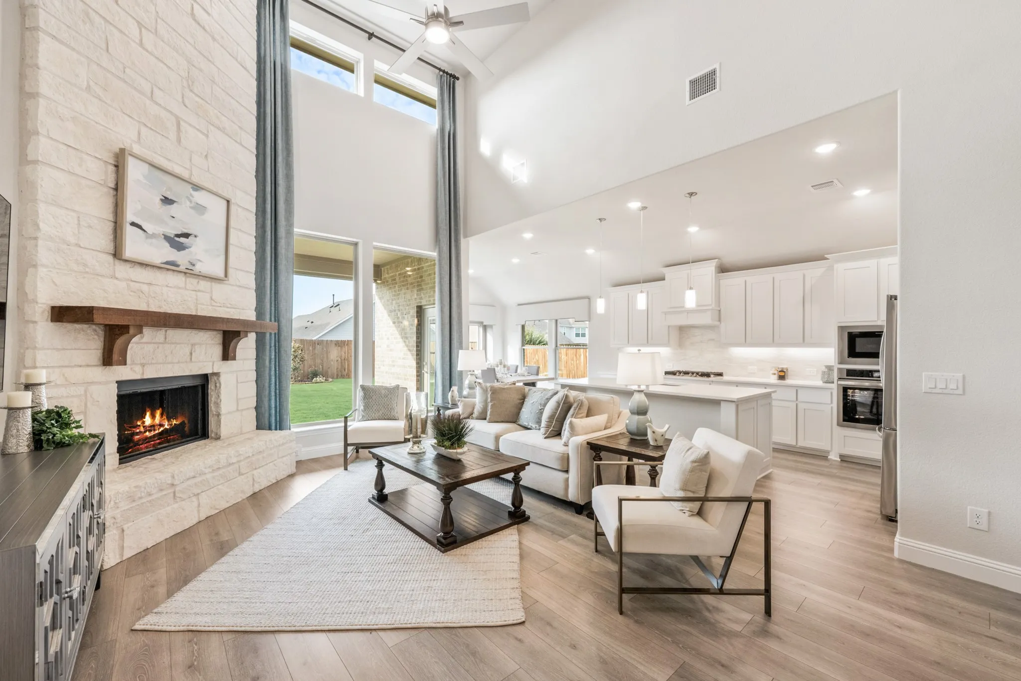 Vaulted ceiling with a ceiling fan, stone fireplace with a wood mantle, large windows, wood-finish flooring, and a kitchen with white cabinetry and stainless steel appliances