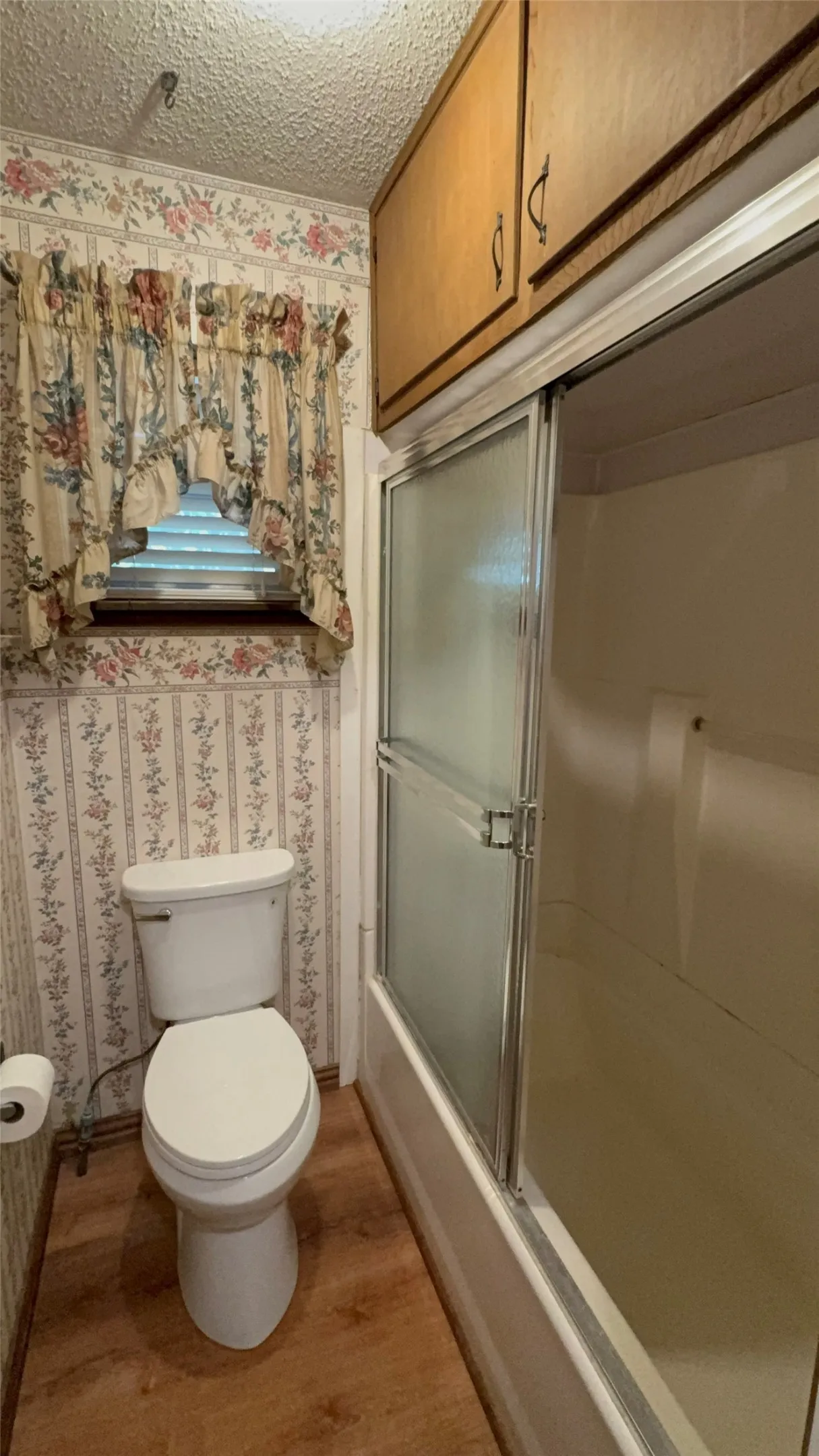 Hallway bathroom. Wood-finish flooring with a toilet, a full bathtub with sliding frosted glass doors, and upper wood cabinetry.