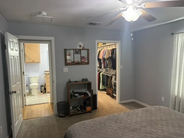 Barndominium - Ceiling fan with light fixture, light gray wall paint, carpet flooring, open closet with built-in organization, bathroom with wood-finish flooring.