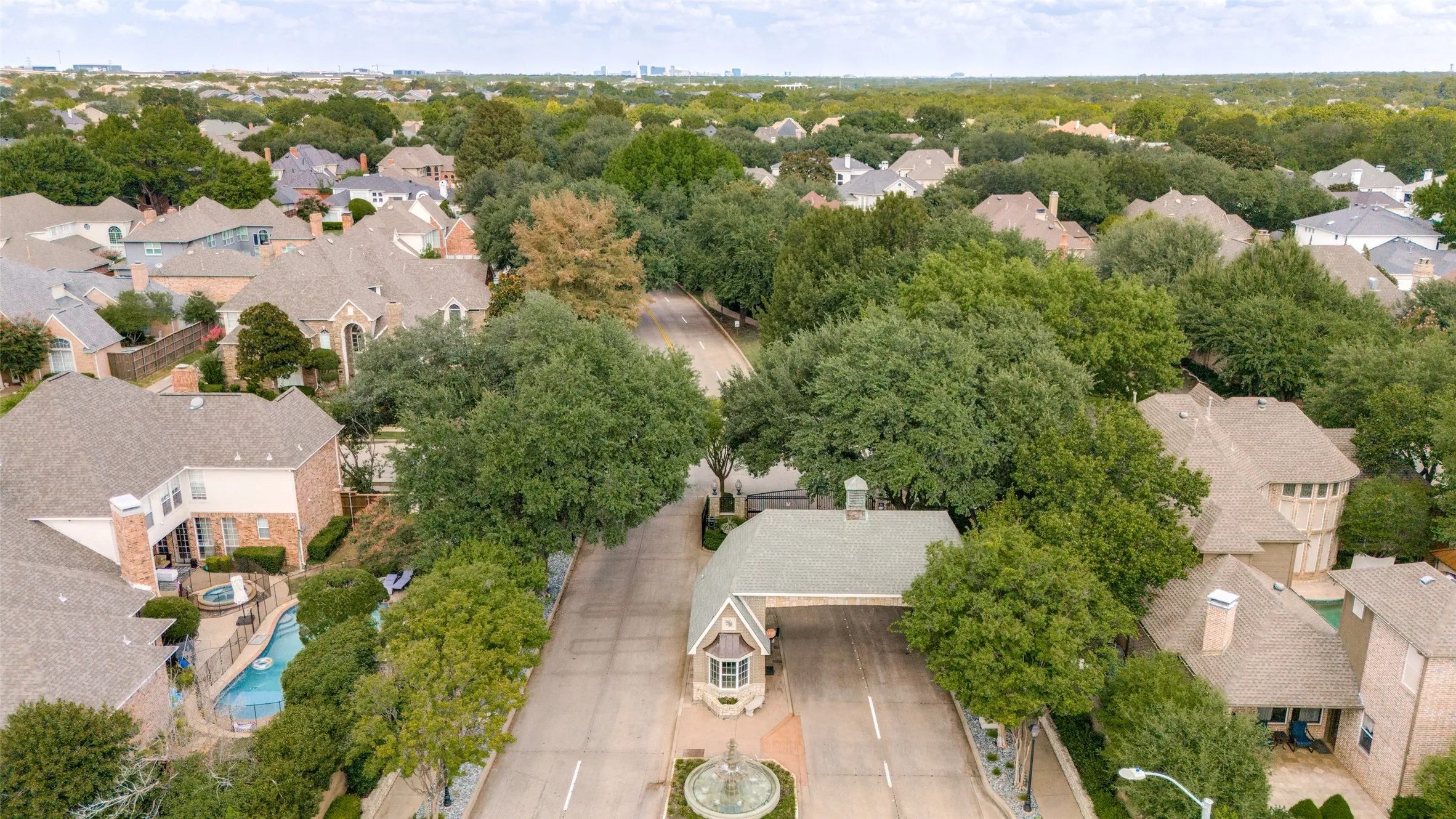 Gated & guarded community entrance featuring a central fountain, mature trees, and a collection of residential properties with varied rooflines
