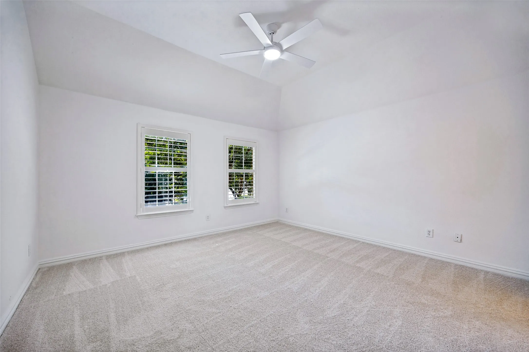 Vaulted ceiling, ceiling fan, two windows with louvered shutters, light-toned carpeting, and white wall finishes