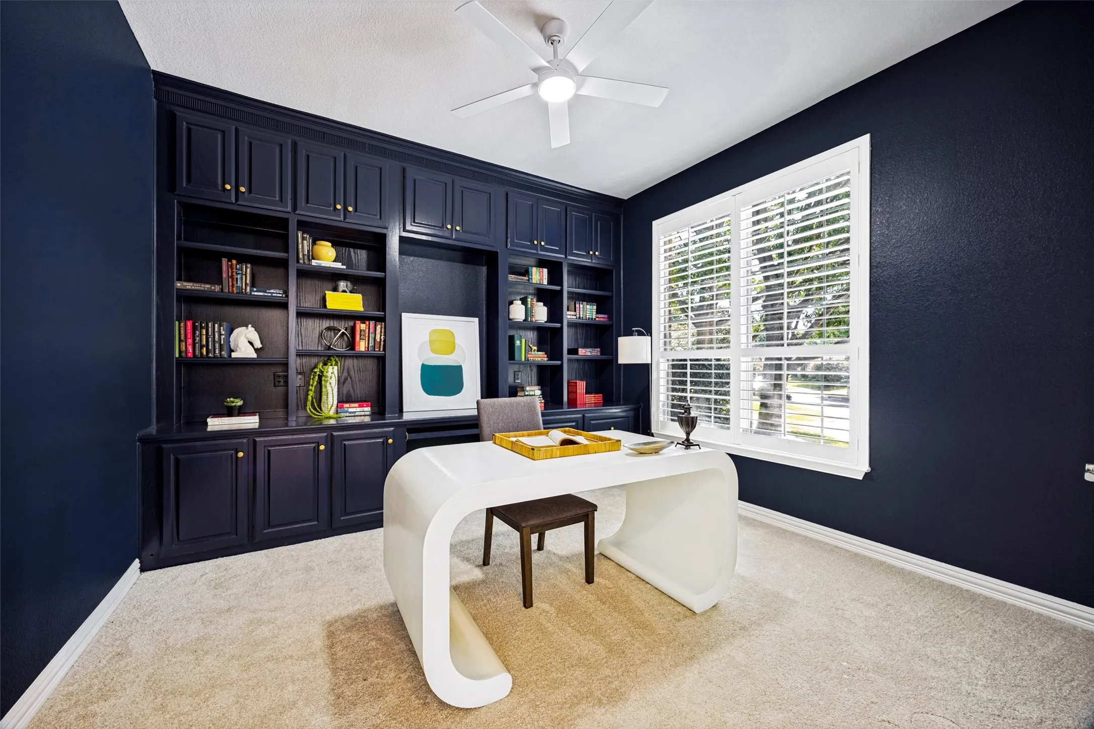Built-in cabinetry with integrated shelving, ceiling fan, window with plantation shutters, and light-colored carpeting