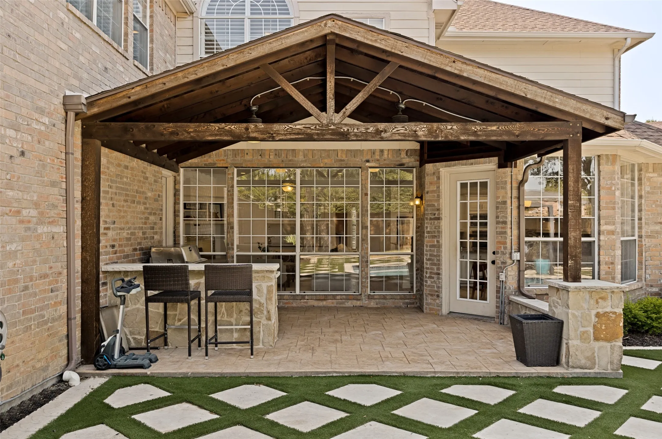 Covered outdoor patio with exposed beam ceiling, built-in outdoor kitchen, stone accent pillars, full-lite French door, and a patterned hardscape