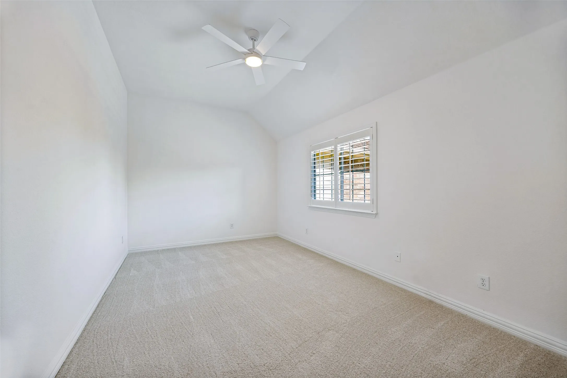 Vaulted ceiling, white ceiling fan with integrated lighting, window with white plantation shutters, light-colored carpeting, and white baseboards