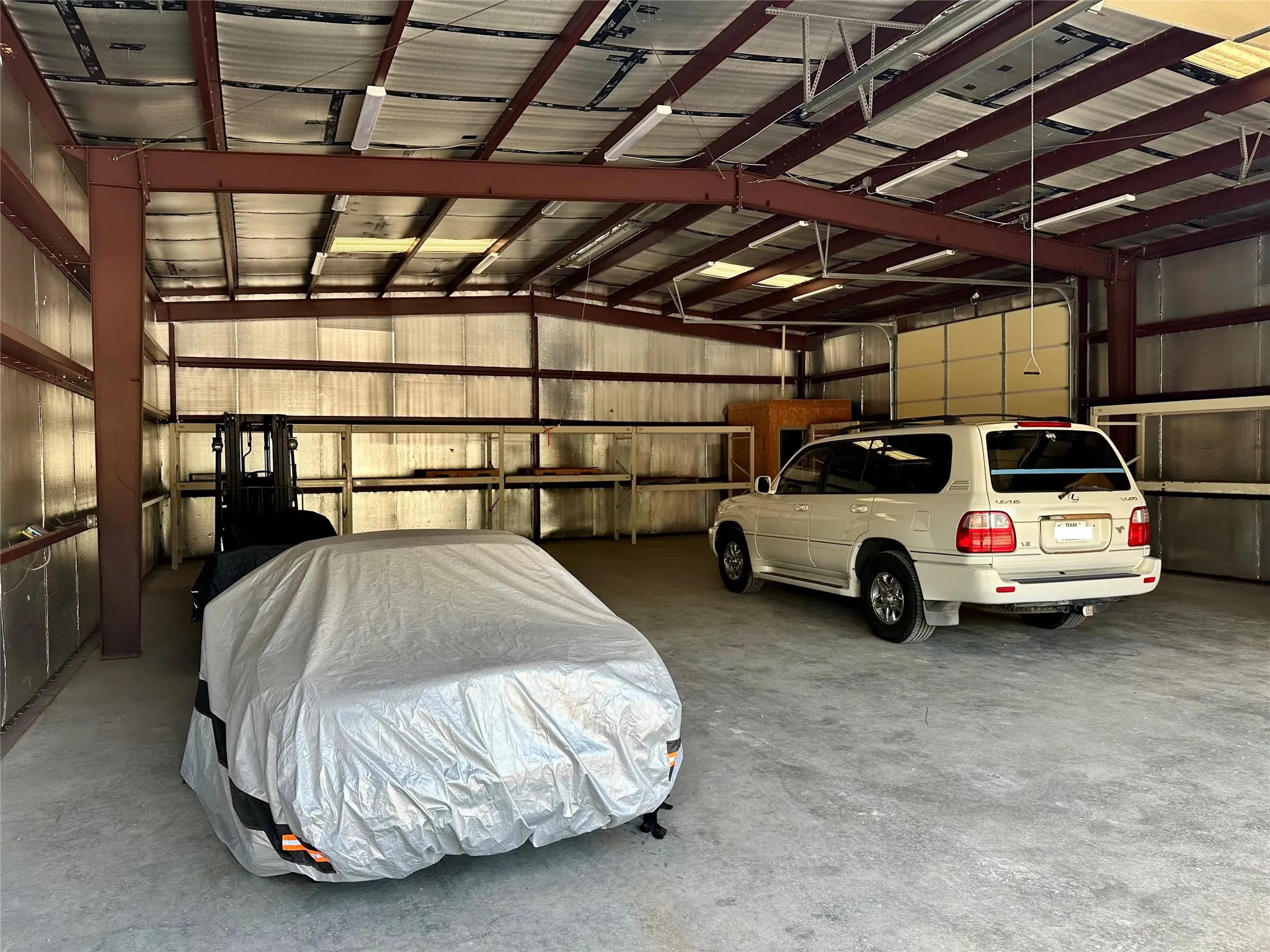 Spacious warehouse featuring a concrete floor, exposed metal beam ceiling structure, fluorescent lighting, and a roll-up garage door