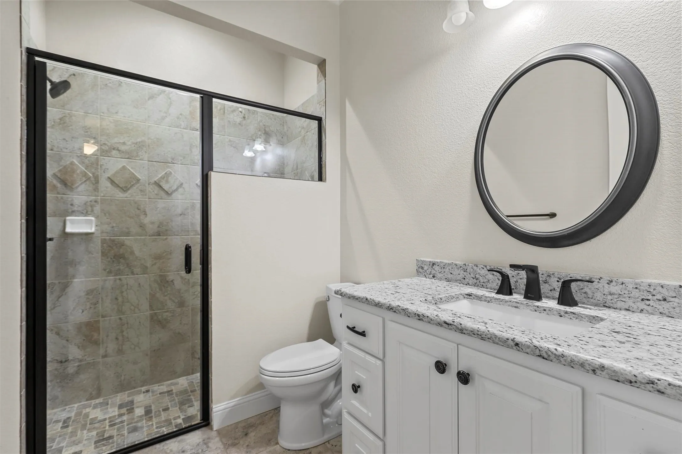 Glass-enclosed shower with tiled walls and floor, white vanity with stone countertop, matte black fixtures, and a circular framed mirror