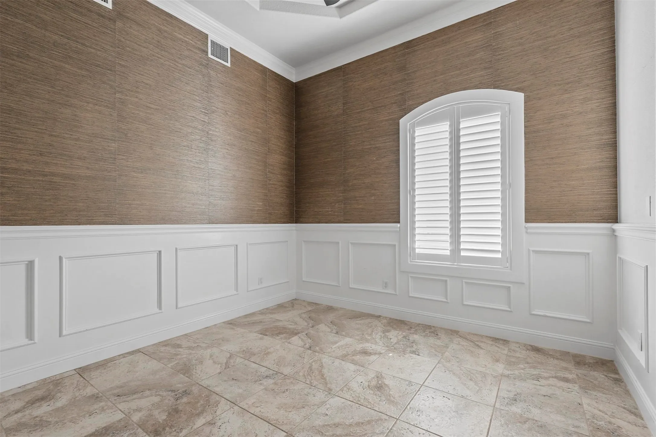 Textured wall covering, crown molding, wainscoting, arched window with plantation shutters, and large format tile flooring