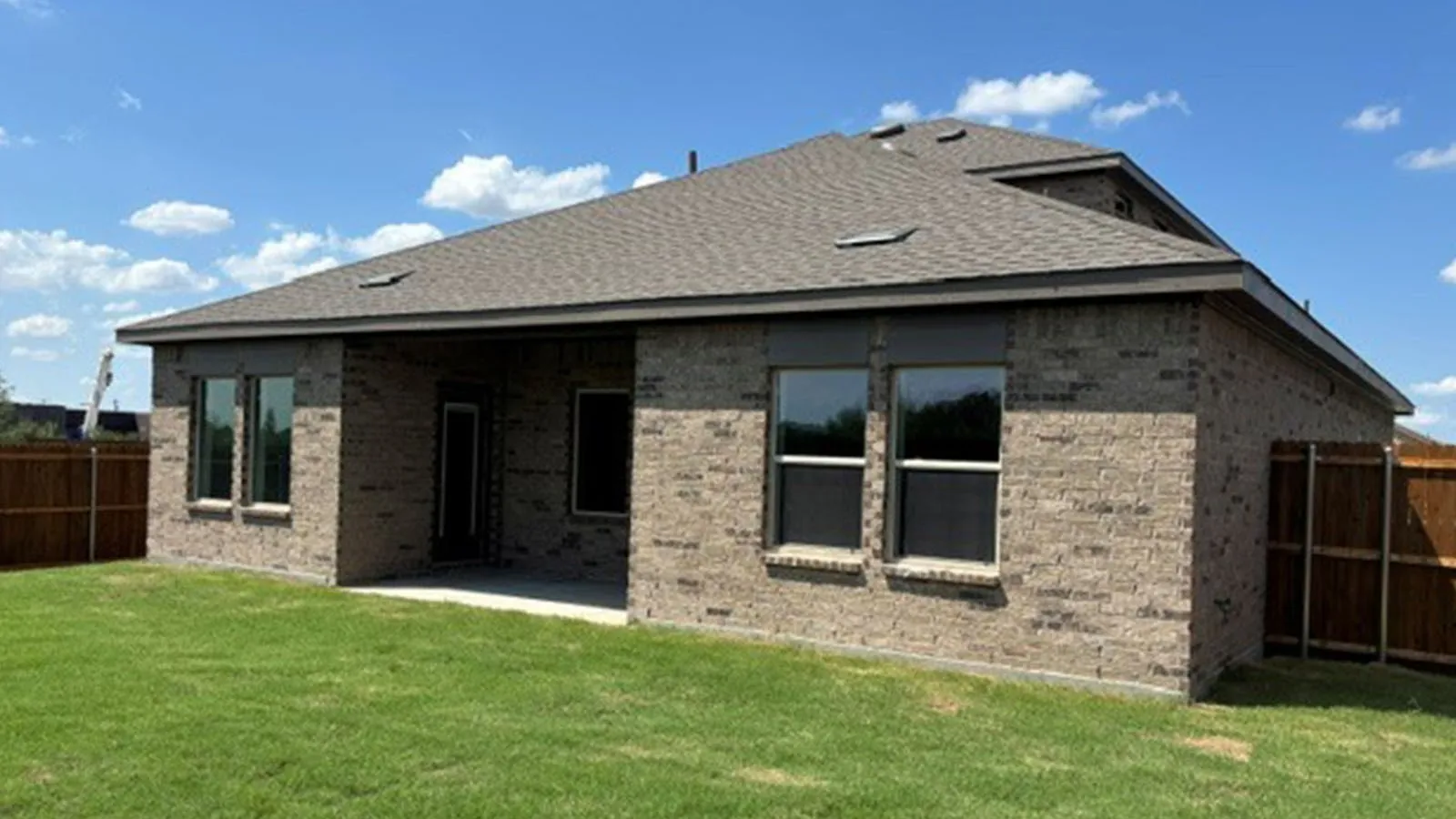 Brick exterior with multiple windows, covered patio, dark shingle roof, and wood fencing