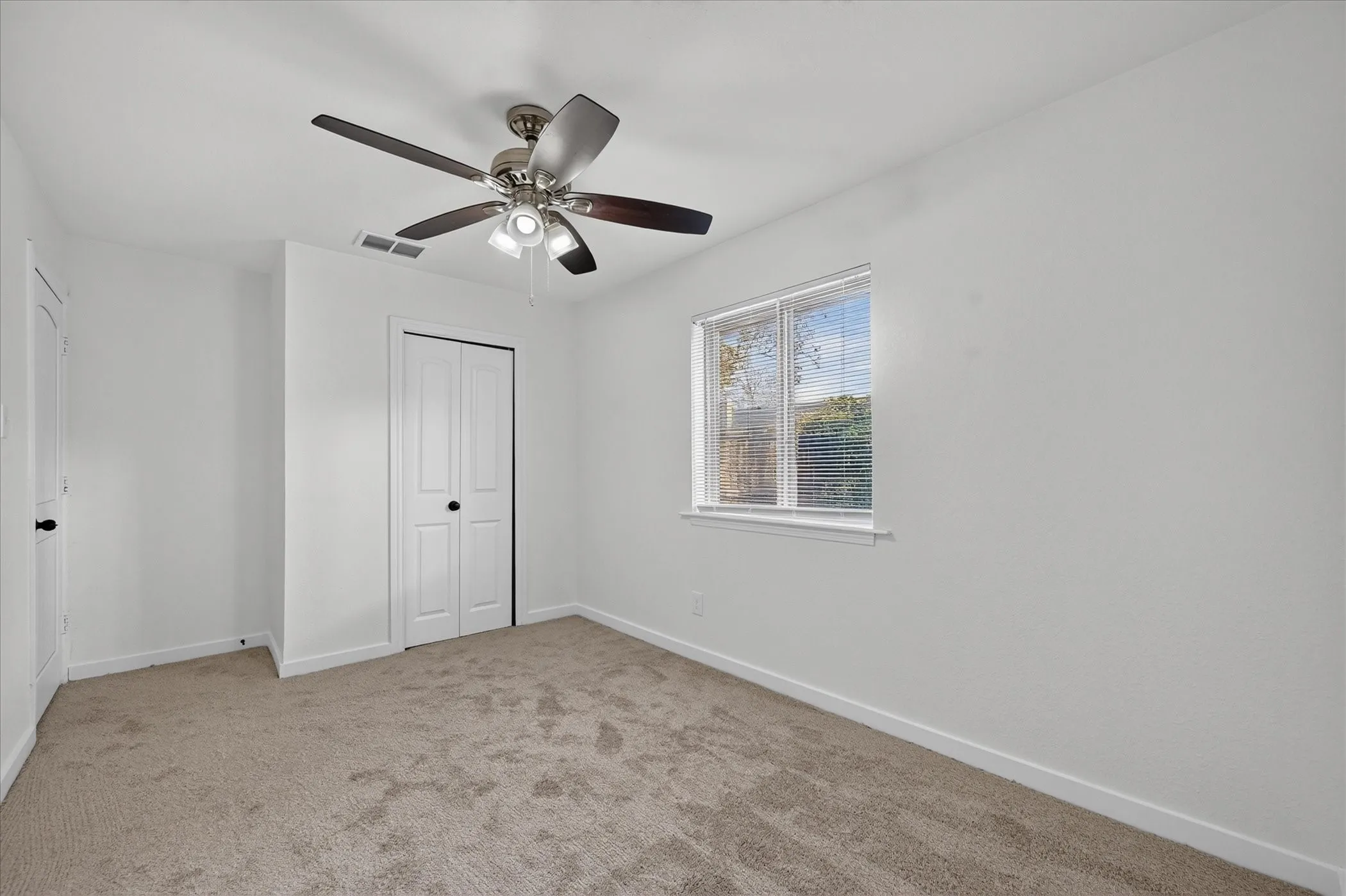 Neutral wall paint throughout, plush carpeting, a multi-pane window with blinds, and a ceiling fan with integrated lighting