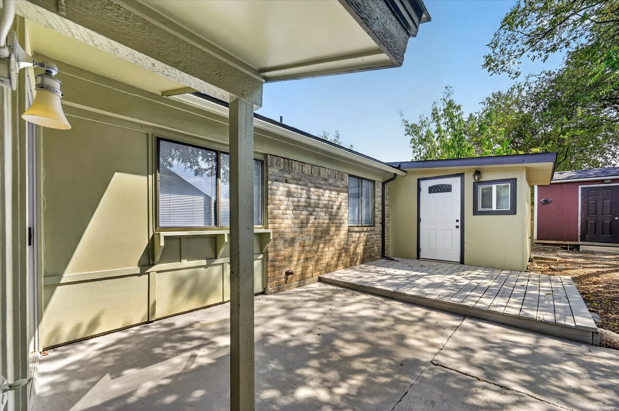 Covered concrete patio with an attached wood deck, exterior wall-mounted light fixture, and a separate structure featuring a white door with an arched window