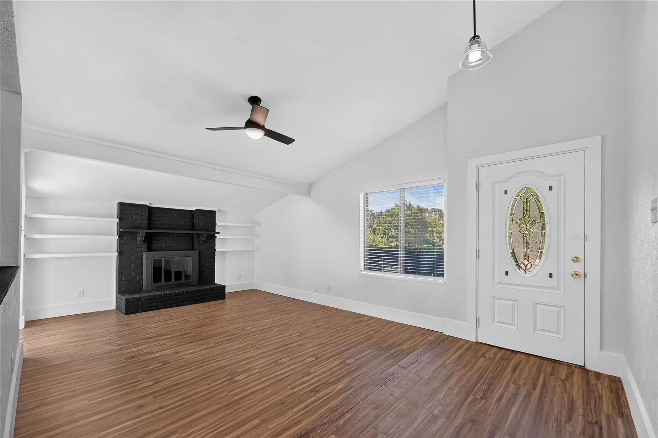 Vaulted ceiling, wood-finish flooring, black brick fireplace, built-in shelving, and a decorative glass front door