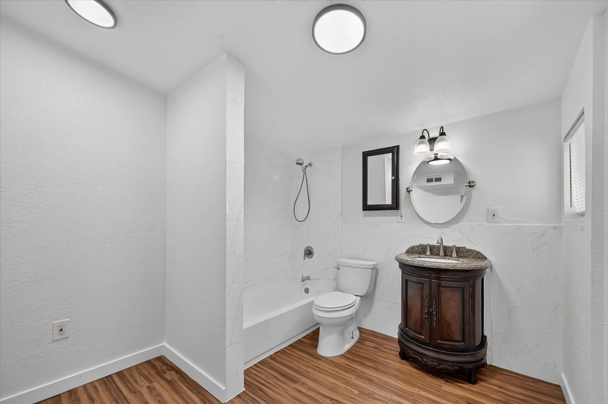 Wood-finish flooring, white walls, built-in bathtub with shower, dark wood vanity with counter, and a circular mirror