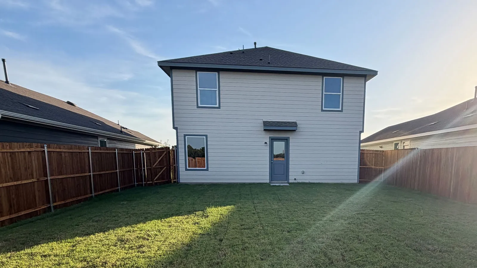 Expansive green lawn, dark wood privacy fencing, two-story exterior with horizontal siding, dark asphalt shingle roof, and a rear entry door with an awning