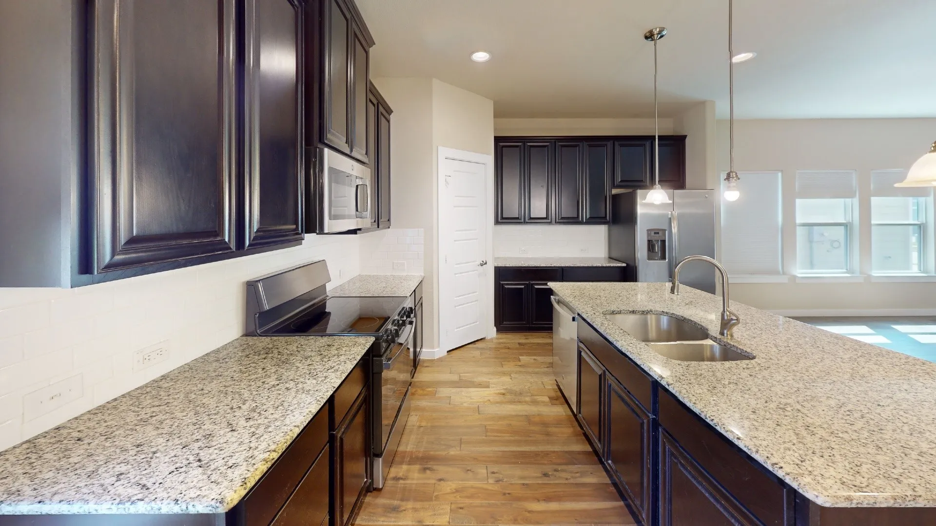 Kitchen featuring dark wood-finish cabinetry, light-toned countertops, stainless steel appliances, a center island with a double sink, and wood-finish flooring
