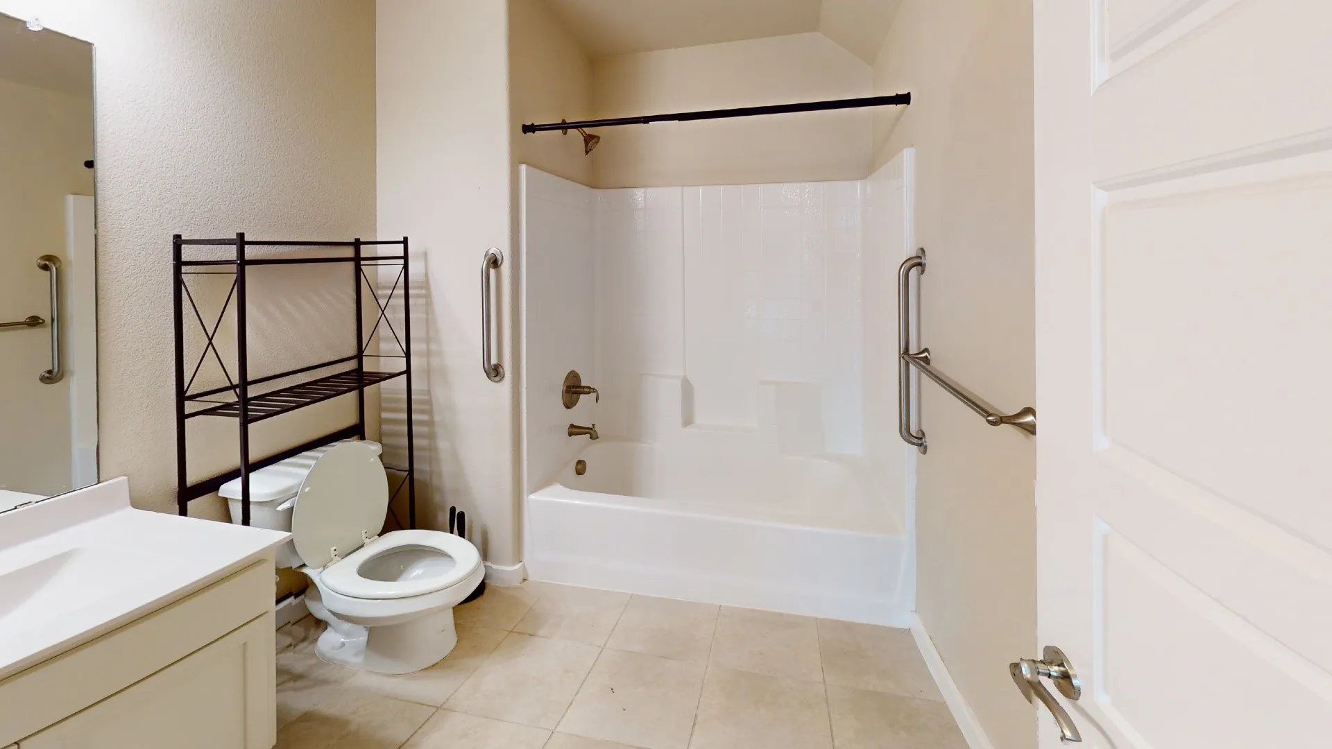 Full-size bathroom featuring a bathtub with an integrated shower, wall-mounted grab bars, a single vanity with a solid countertop, and tile flooring