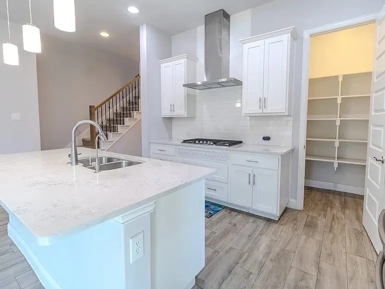 Kitchen featuring an island with an integrated sink, white cabinetry, a gas range with a stainless steel range hood, subway tile backsplash, and a built-in pantry