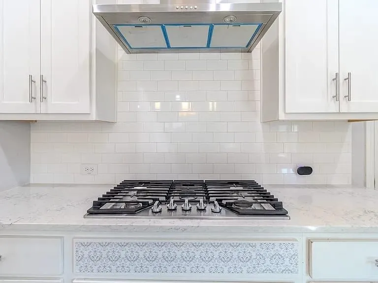 Stainless steel gas range with a coordinating hood, white subway tile backsplash, white shaker-style cabinetry, and light-toned countertops