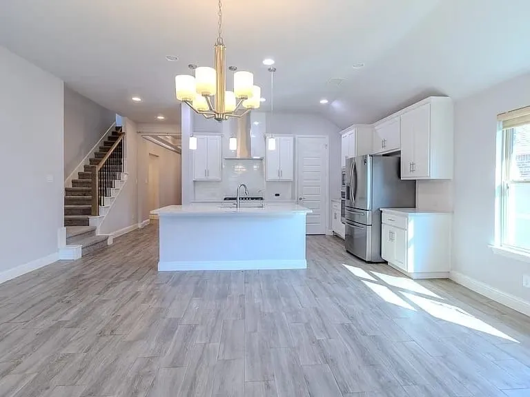 Spacious open-concept living area featuring wood-look tile flooring, a kitchen island with seating, stainless steel appliances, white cabinetry, and a carpeted staircase with a metal railing